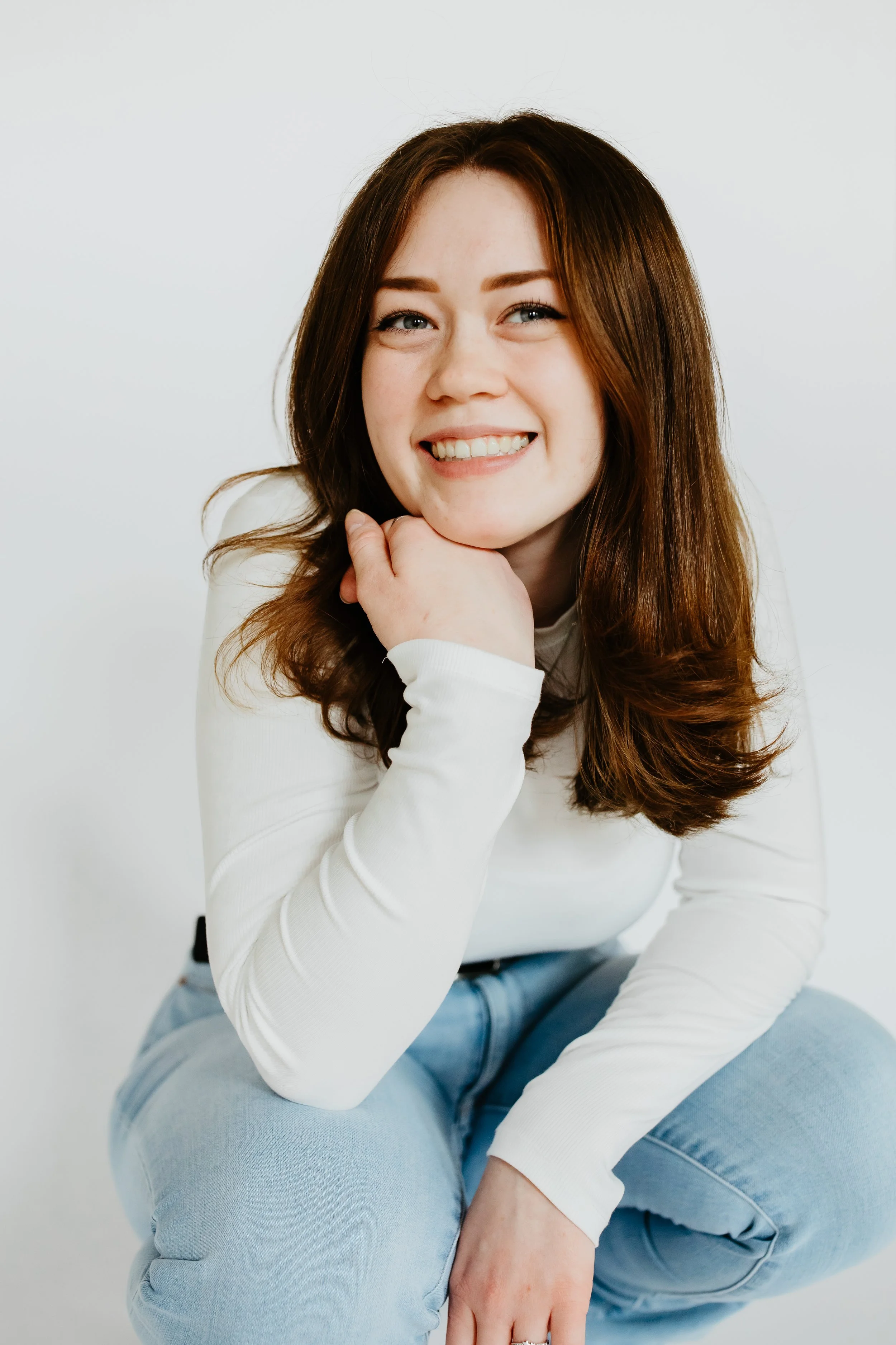 A woman with reddish-brown hair smiling, sitting with her chin resting on her hand against a white background.