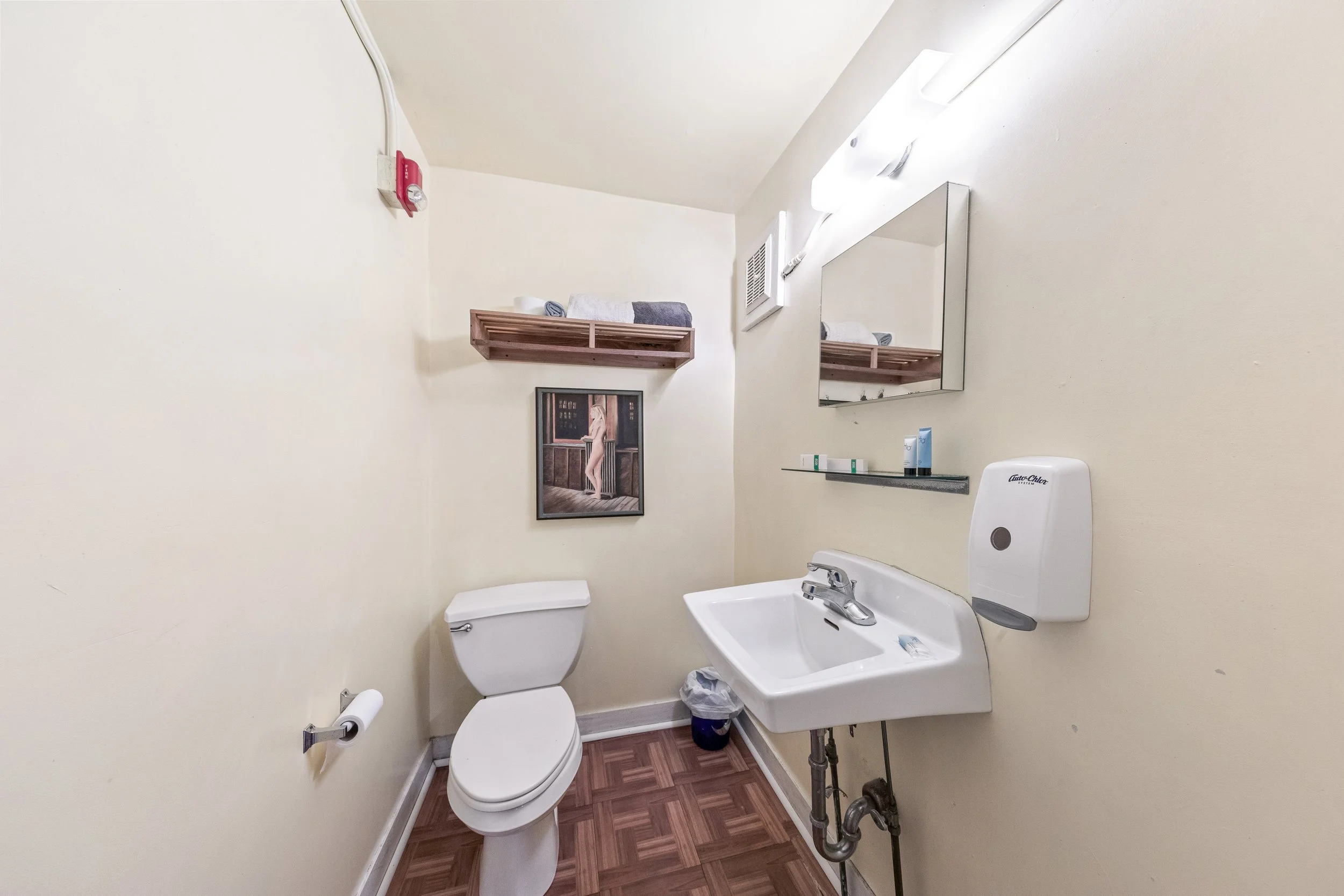 Small bathroom with a white toilet, a wall-mounted sink, a mirror, wooden shelf with towels, a framed picture, a soap dispenser, and toiletries on a glass shelf.