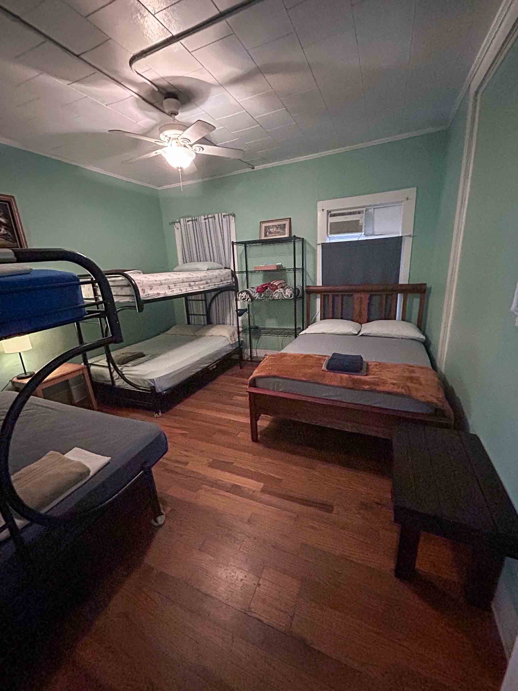 A bedroom with mint green walls, a ceiling fan, a bunk bed, a double bed with a folded blanket, a small end table with a lamp, a black shelving unit with blankets, and two windows with curtains or shades.