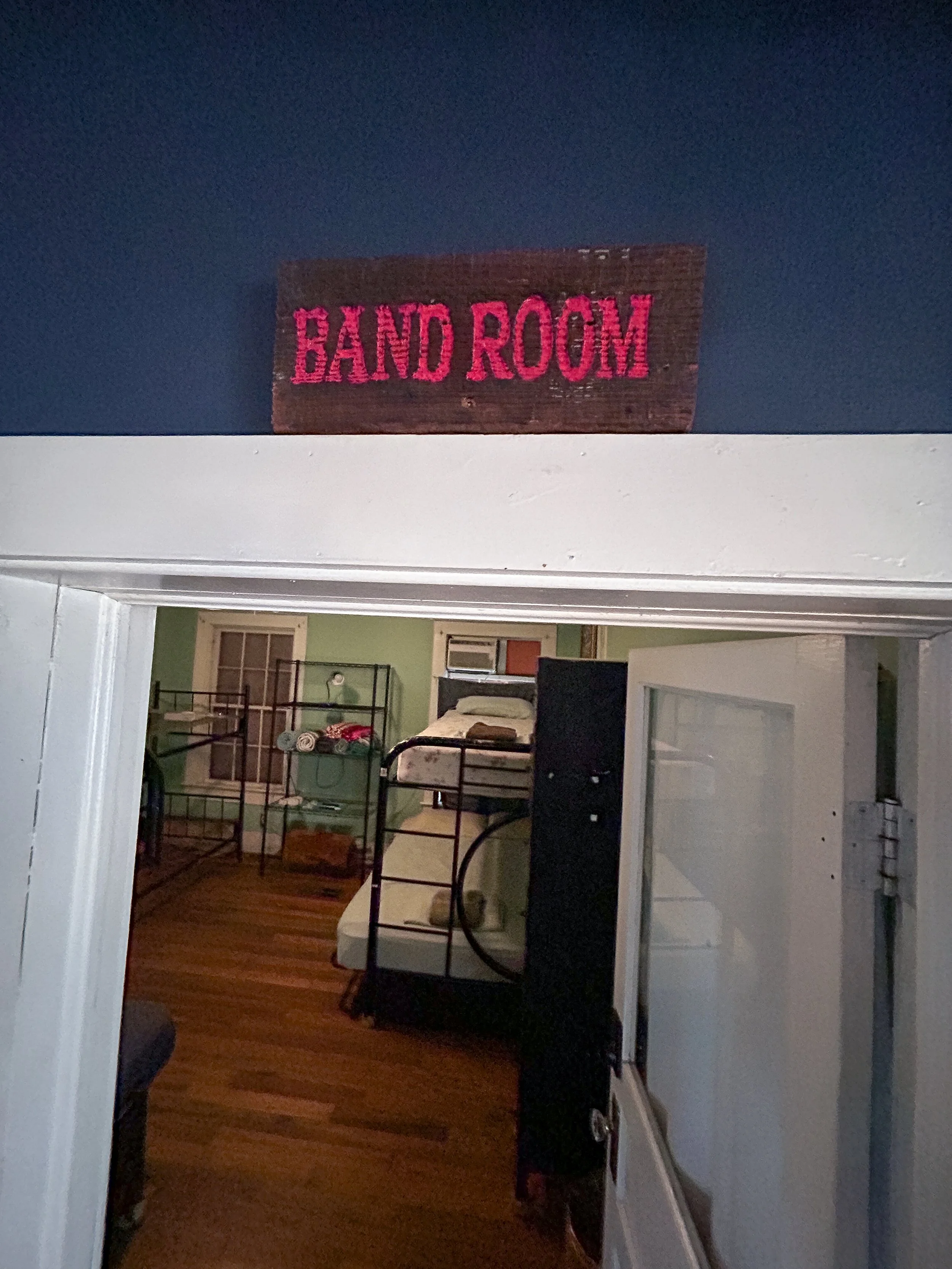 View through a doorway into a room with bunk beds and a window. A sign above the doorway reads 'BAND ROOM' in pink text on a wooden background.