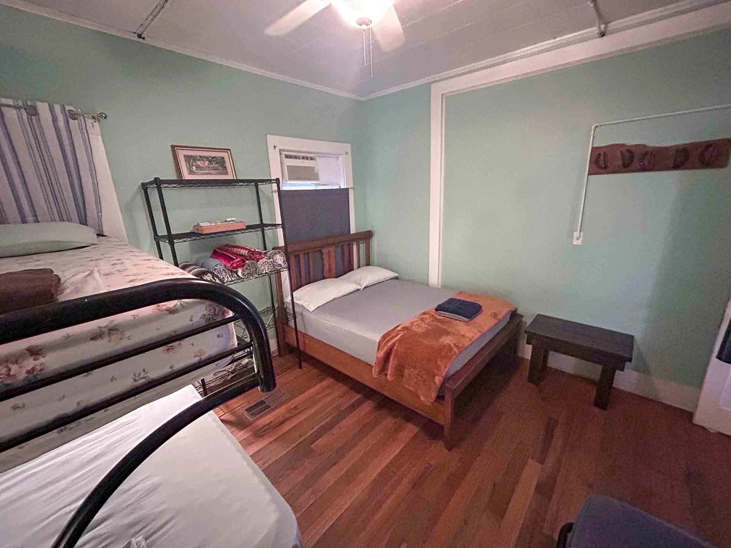 A bedroom with two beds, one with a floral pattern and the other plain, with towels on top, a black metal bookshelf with folded towels and picture frames, a small wooden side table, green walls, and a window with blinds.