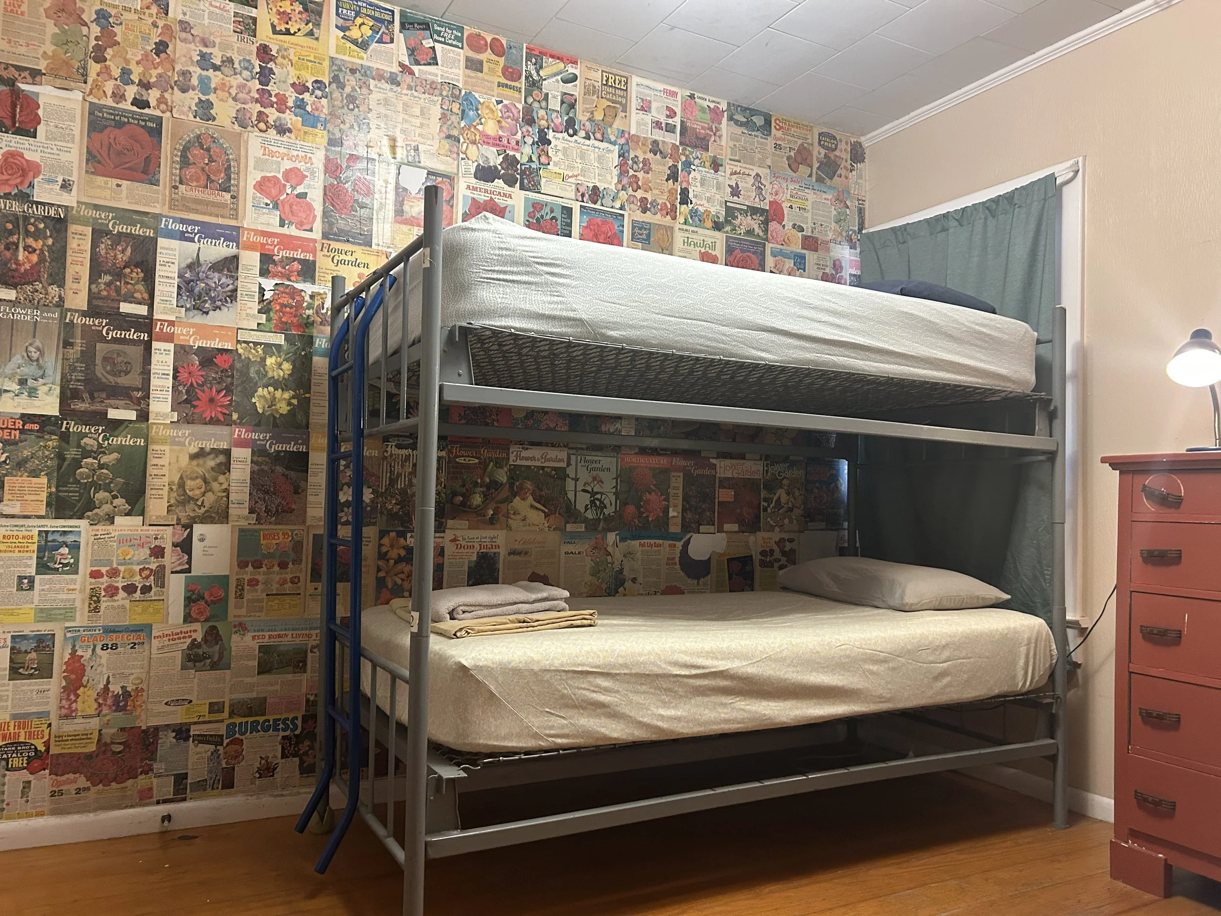 A bedroom featuring a metal bunk bed with a mattress on top and a mattress on the bottom, pillows on the beds, and a wall decorated with vintage flower and gardening magazine covers. There is a small red dresser with drawers and a lamp on it on the right side of the image.