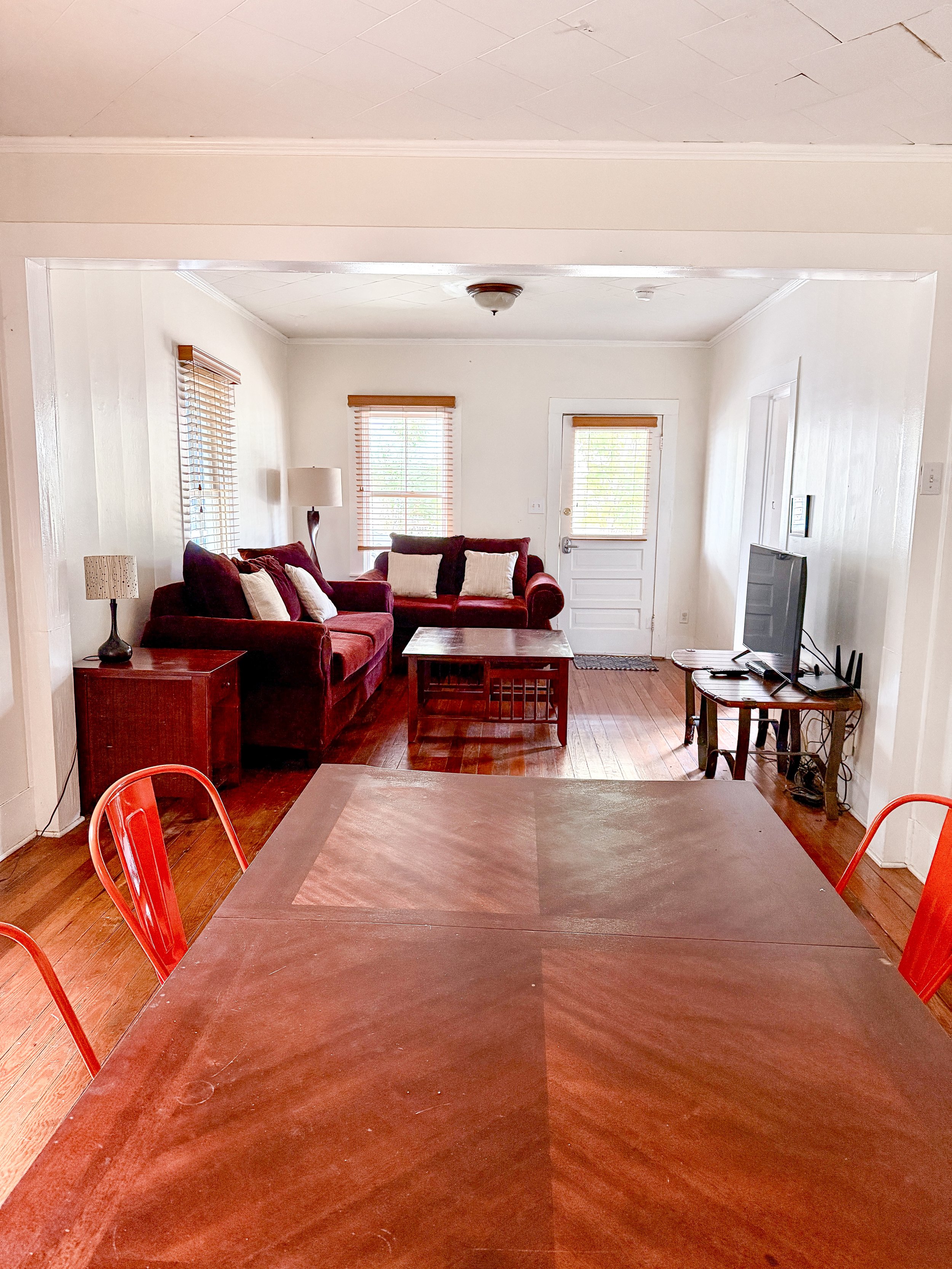 Living room with two red sofas, a wooden coffee table, a TV on a small wooden stand, and three windows with blinds. A door with a window in the background, and a ceiling light fixture.