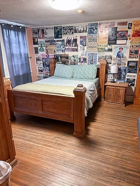 A bedroom with a wooden bed frame, a wall decorated with vintage posters, a nightstand with a lamp, and hardwood floors.