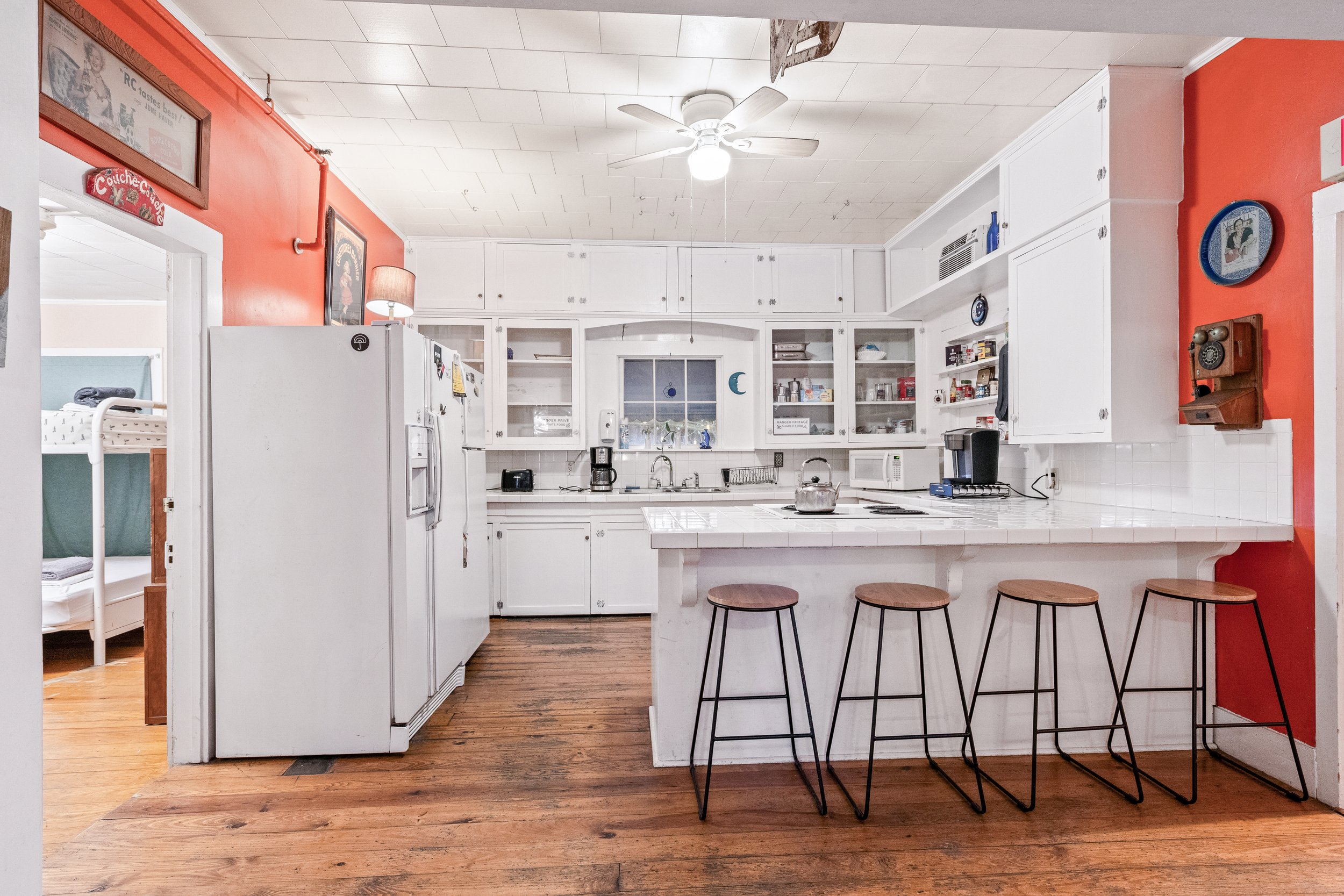 A kitchen with white cabinets, a white tile countertop, and a small window above the sink. Four wooden barstools with black metal legs are placed around a white tiled island. The left wall is painted red, and a vintage rotary phone is mounted on it. 