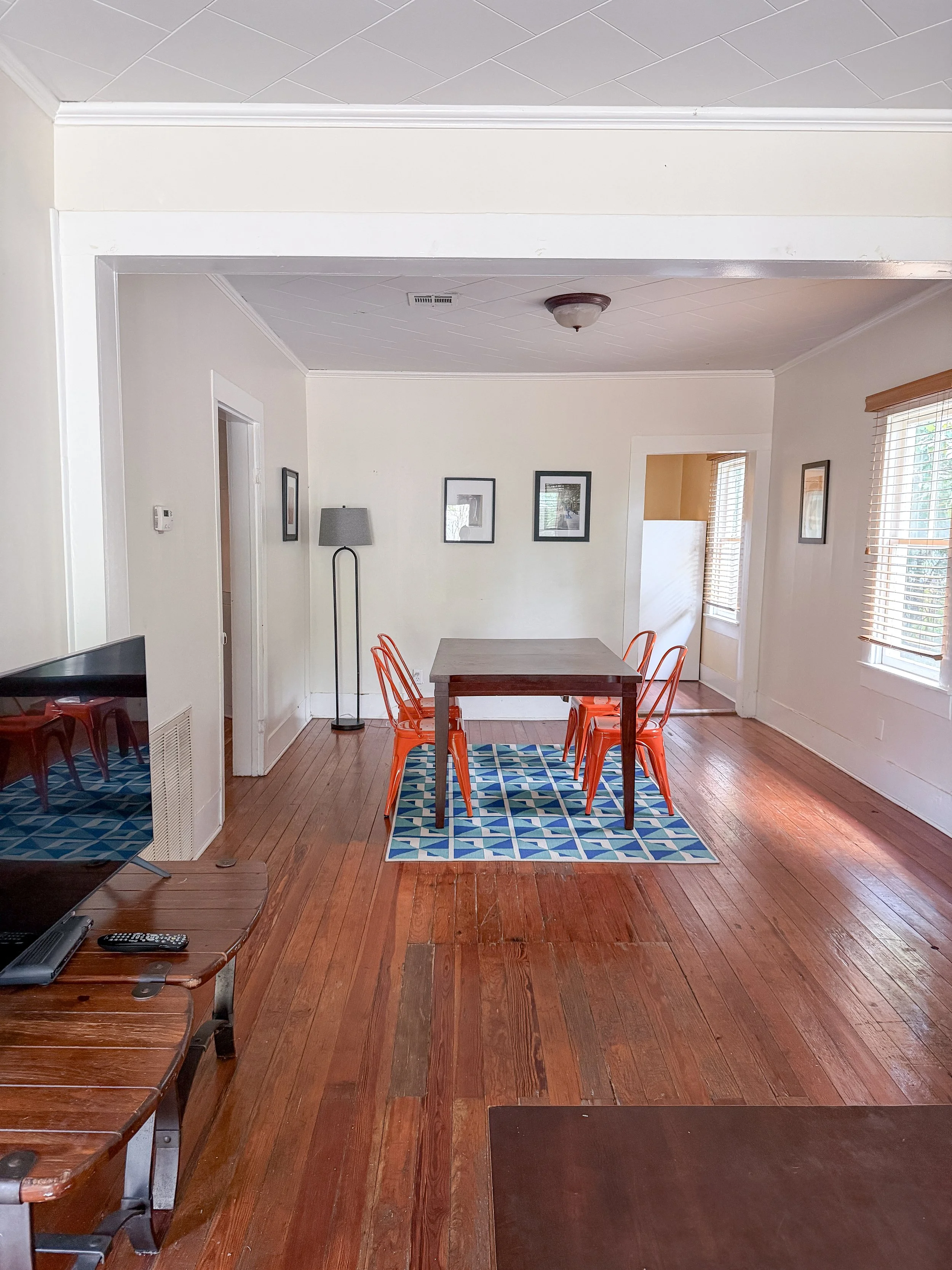 A room with hardwood floors, a dining table with orange chairs, a rug underneath, a standing floor lamp, framed pictures on white walls, and windows with blinds letting in natural light.