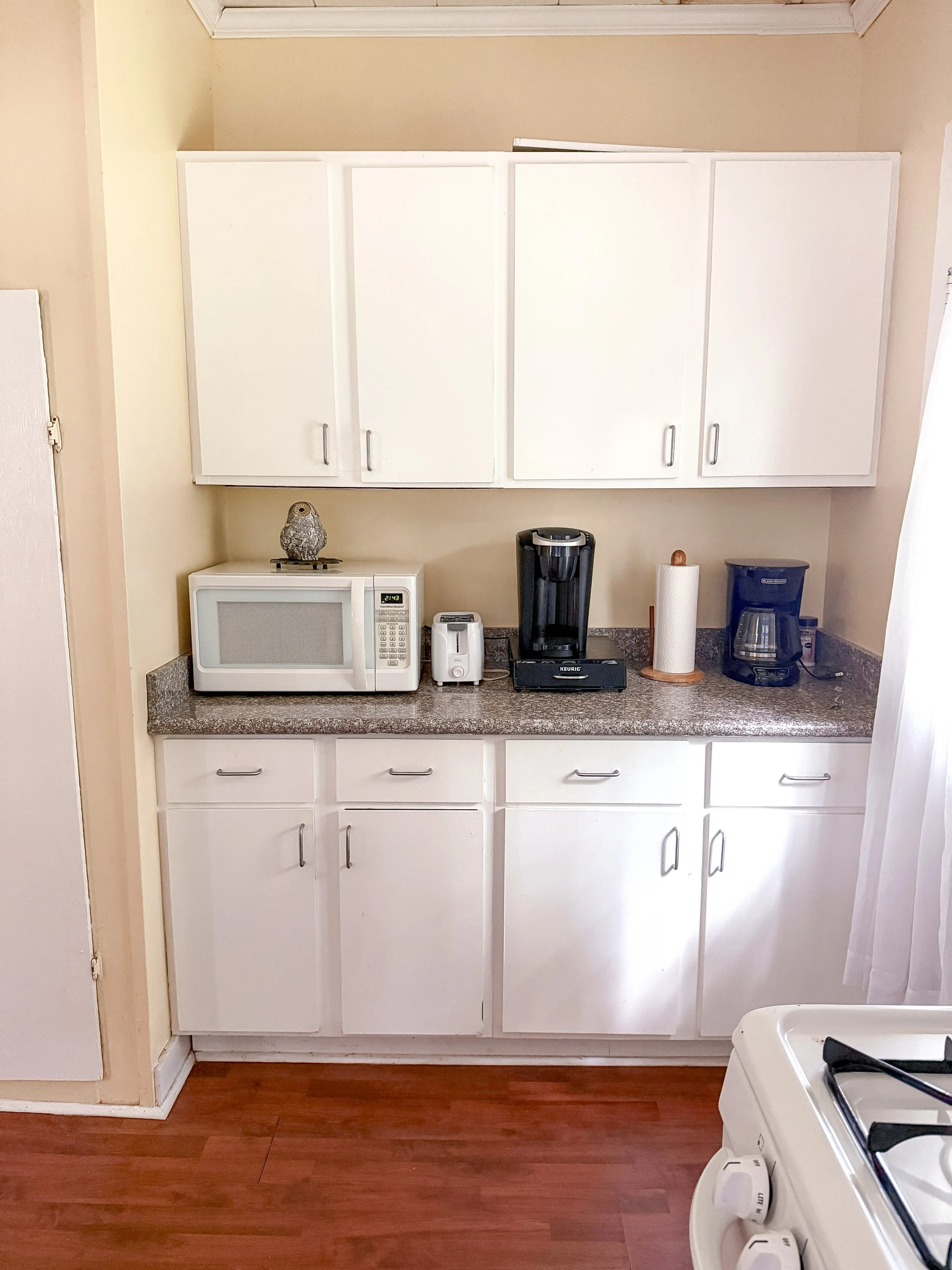 Kitchen countertop with microwave, coffee maker, and paper towel roll, white cabinets above and below, hardwood floor, and stove in the foreground.