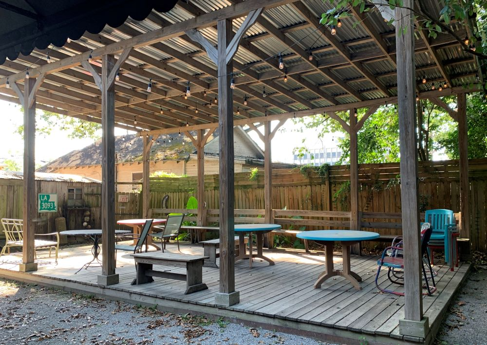 A wooden outdoor patio with a metal roof, string lights, and various chairs and tables, surrounded by a wooden fence and greenery.