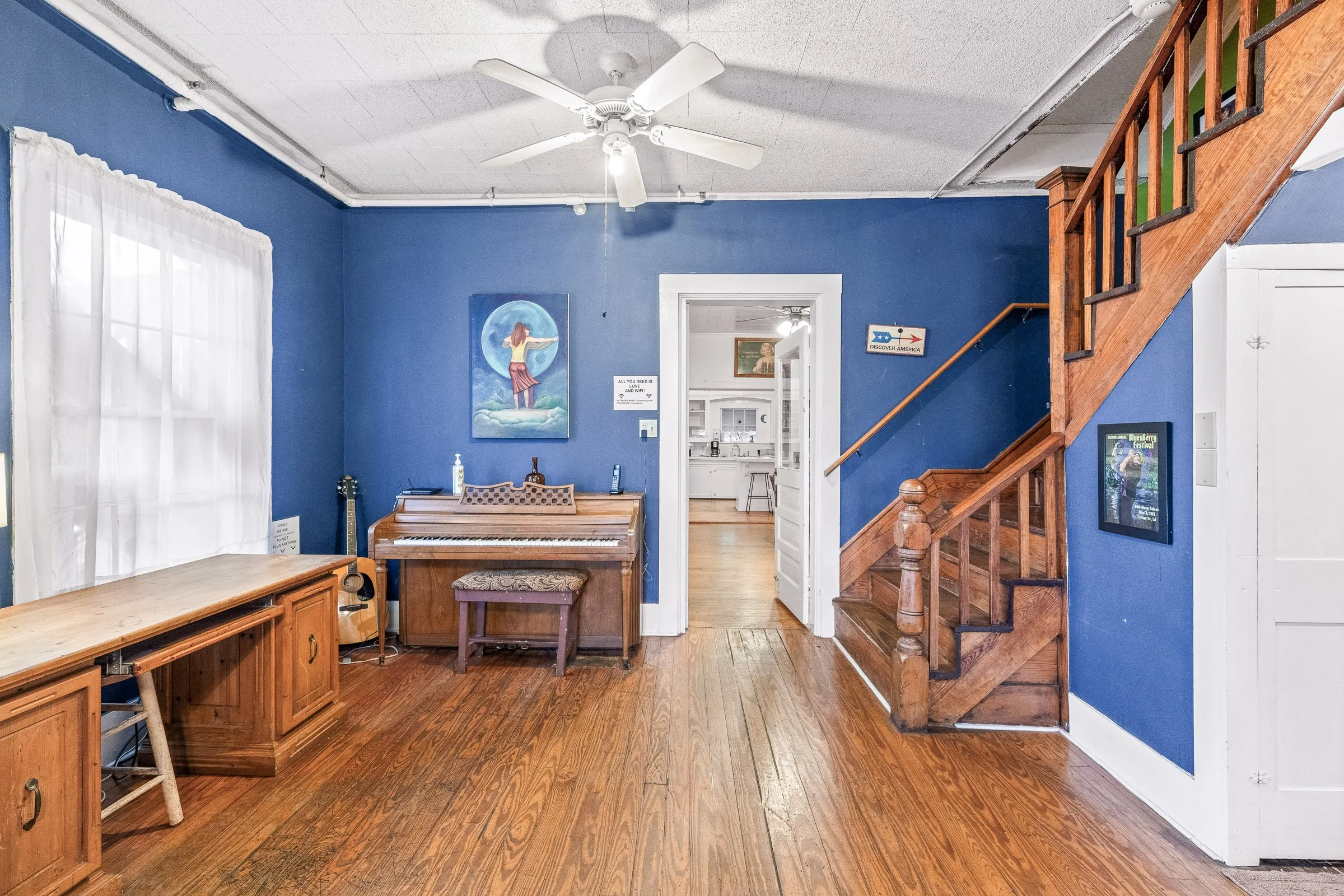 Living room with blue walls, white ceiling fan, wooden staircase, window with white curtains, desk, piano, and framed artwork.