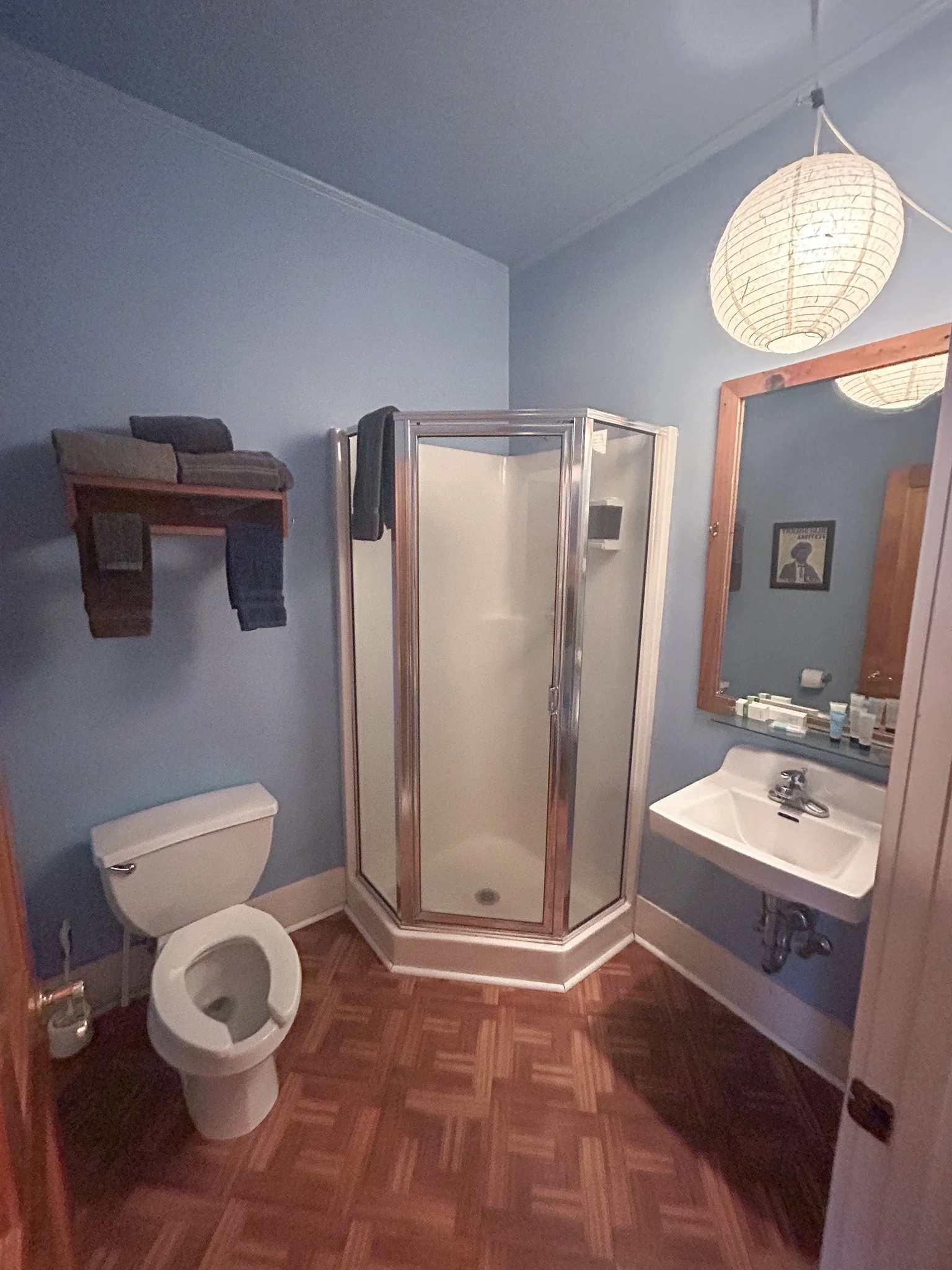 Small bathroom with blue walls, a corner shower with frosted glass doors, a white toilet, a small white sink, a large wooden framed mirror, a ceiling paper lantern, and a wooden shelf holding folded towels.