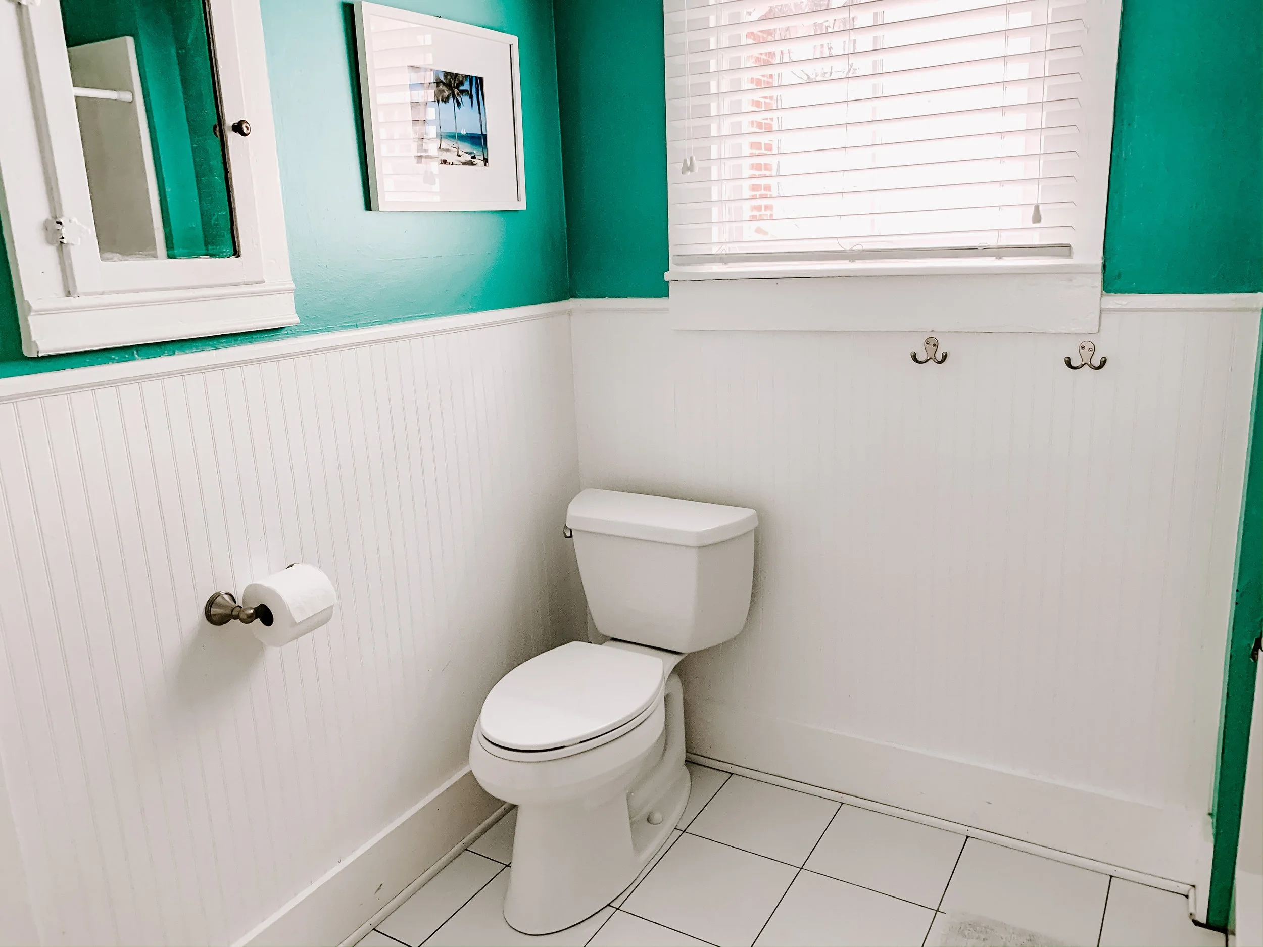 A small bathroom with a white toilet, a white window with blinds, teal upper walls, white beadboard lower walls, and wall hooks. A framed picture of a beach scene hangs on the teal wall.