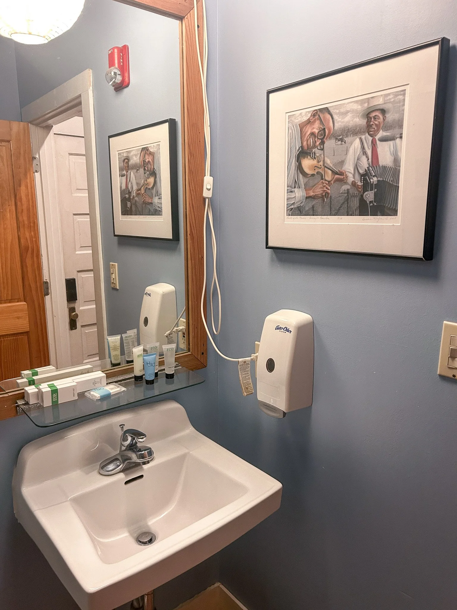 A bathroom sink with a mirror above it, toiletries on a glass shelf, a wall-mounted soap dispenser, and framed artwork of musicians playing instruments, with a door and wooden trim visible.