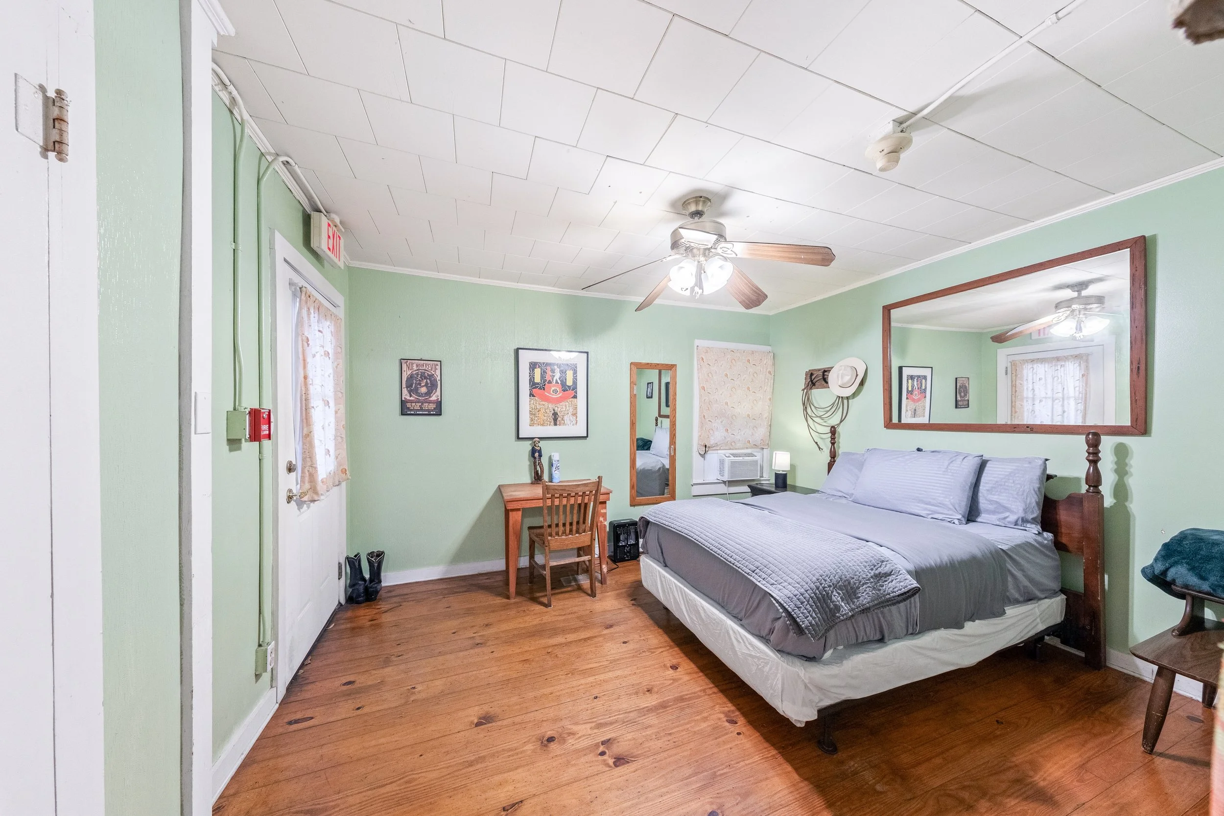 A bedroom with light green walls, wooden floor, a double bed with gray bedding, a large mirror above the bed, a ceiling fan, a window with floral curtains, artwork on the walls, a small wooden desk with a chair, and a mirror on the door.