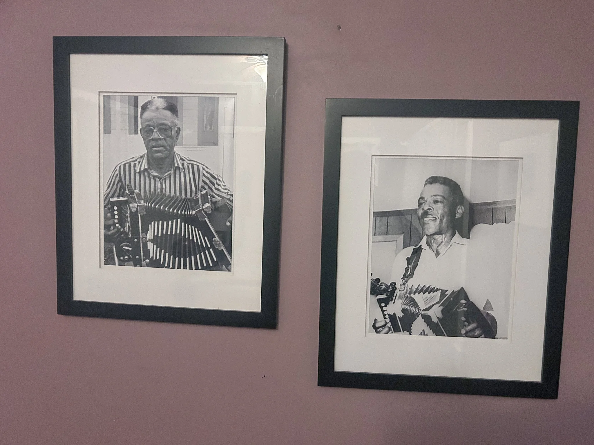Black and white photographs of two men playing musical instruments, framed and hung on a pink wall.