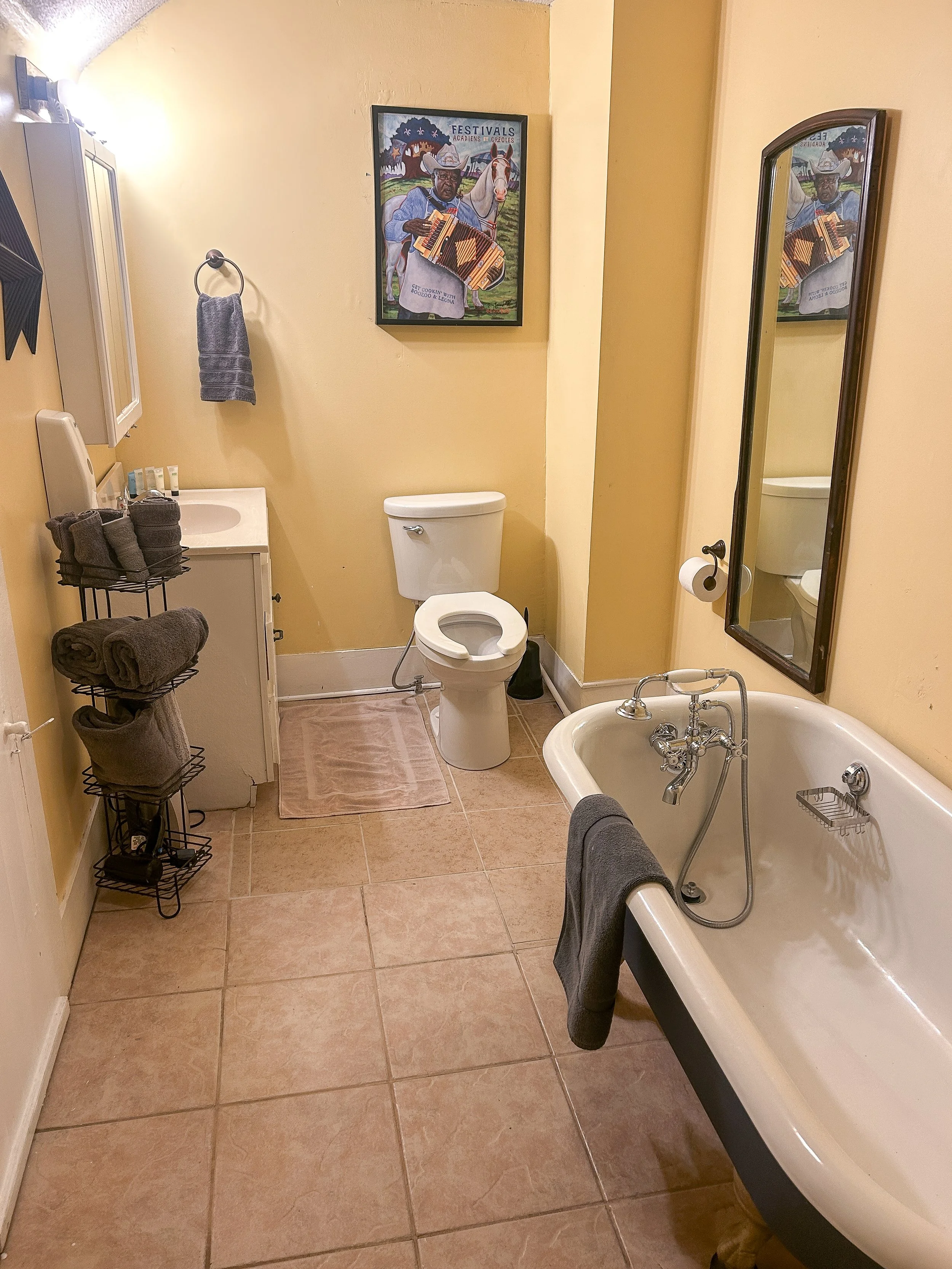 A vintage-style bathroom with yellow walls, a white toilet, a bathtub with a gray towel hanging on the side, a mirror, a small sink with a cabinet, a towel rack with gray towels, a picture of a musician playing a keyboard, and tile flooring.