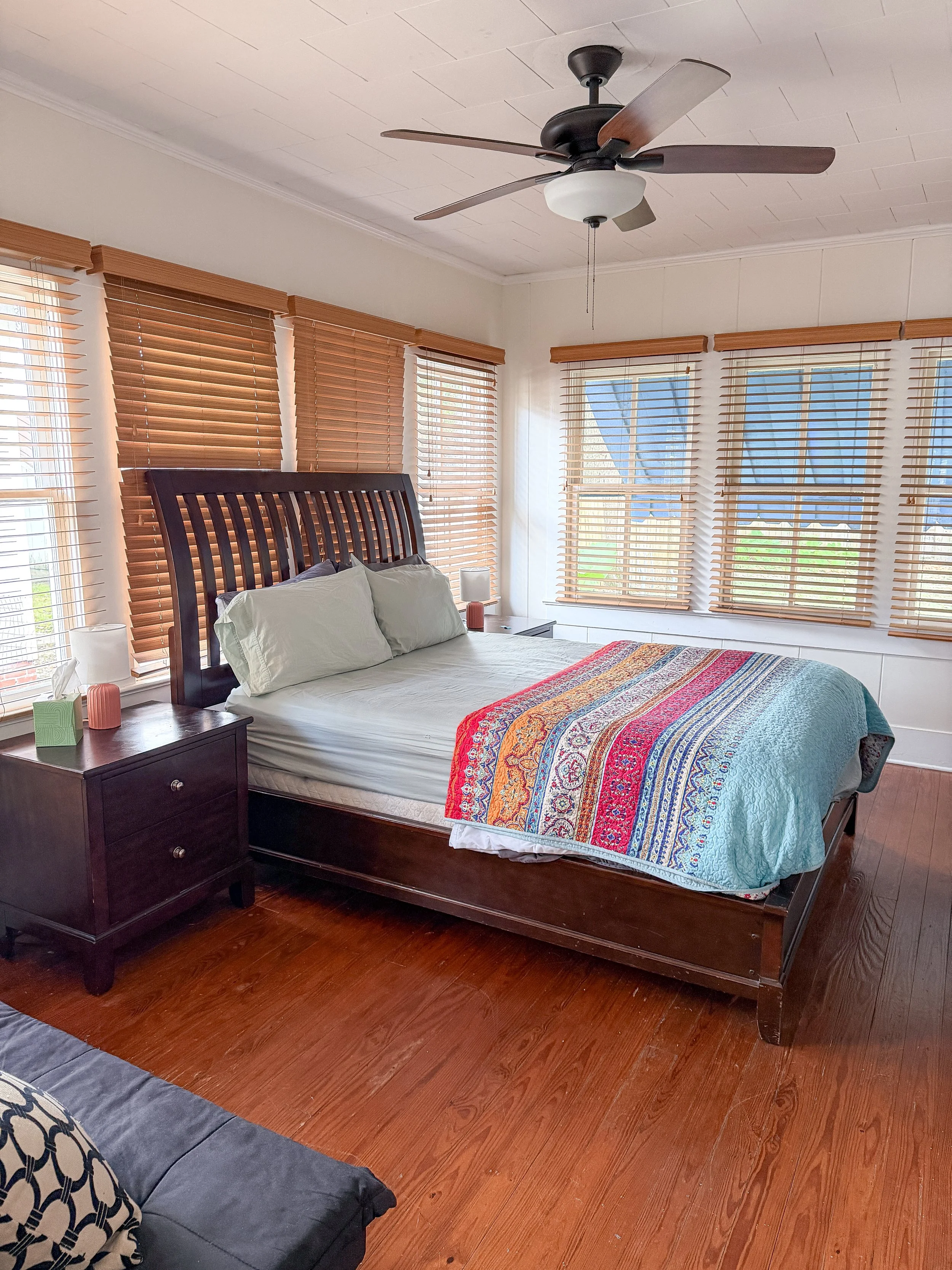 A cozy bedroom with a wooden bed frame, white bedding, colorful quilt, wooden nightstands on either side, the left with a lamp and tissue, the right with a lamp, multiple windows with wooden blinds, ceiling fan, and hardwood floors.