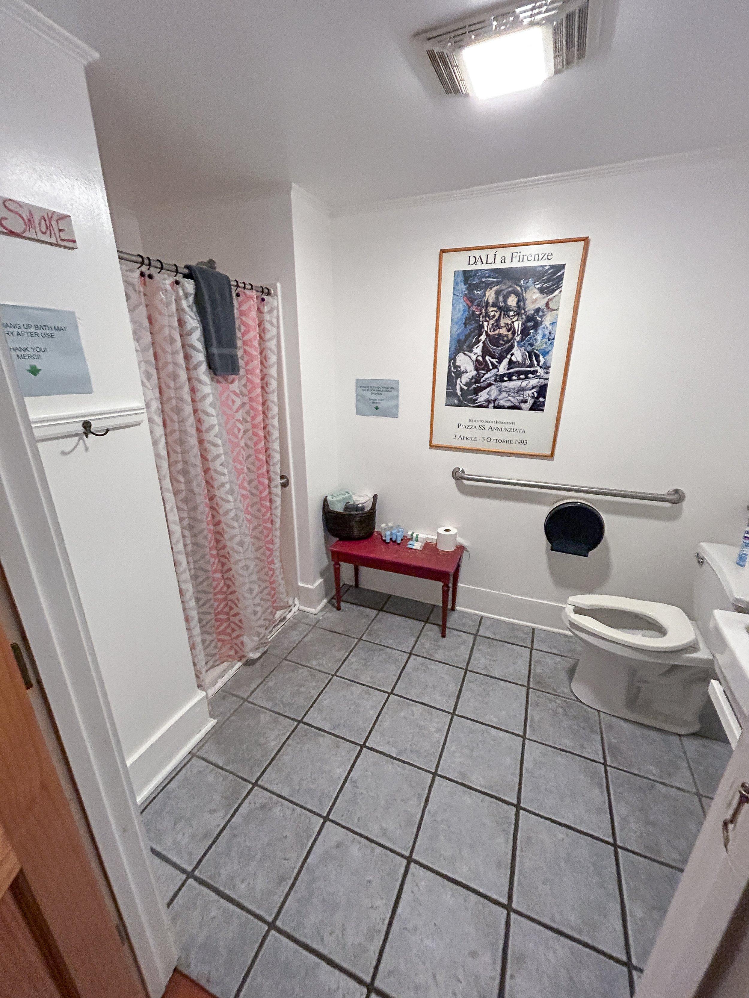 A clean, well-lit bathroom with a tiled floor, a toilet, and a shower area behind a pink and white patterned curtain. There is a small red table with toiletries, a wastebasket, and a roll of toilet paper. A large framed poster of a Salvador Dalí artw