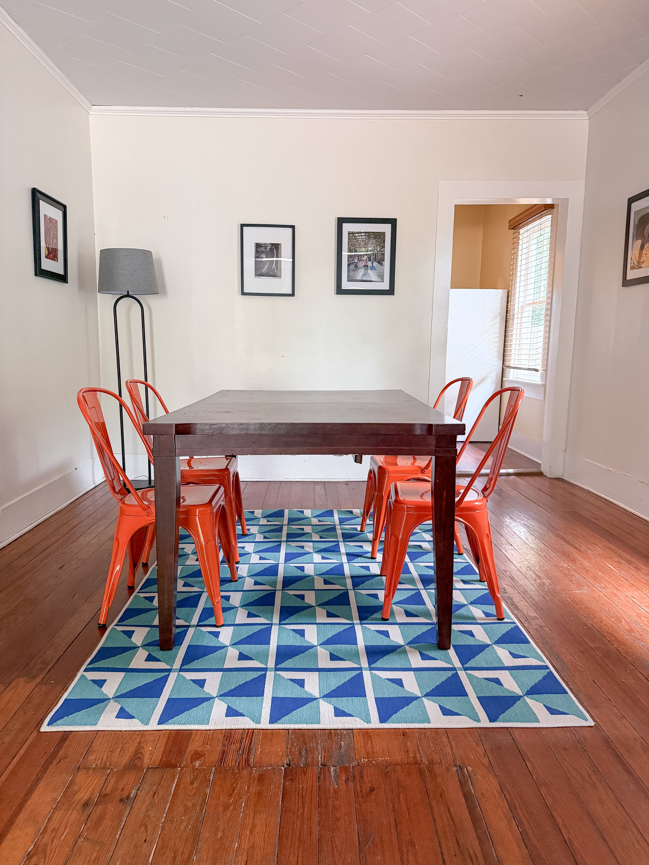 A dining room with a wooden table, six orange metal chairs, a blue patterned rug, framed pictures on the walls, a floor lamp, and a window with shutters.