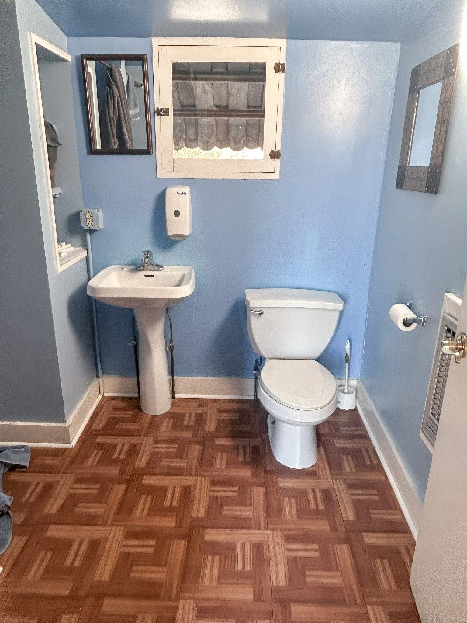 Small bathroom with blue walls, a white toilet, a pedestal sink, a mirror, and a window with a small storage cabinet above it.