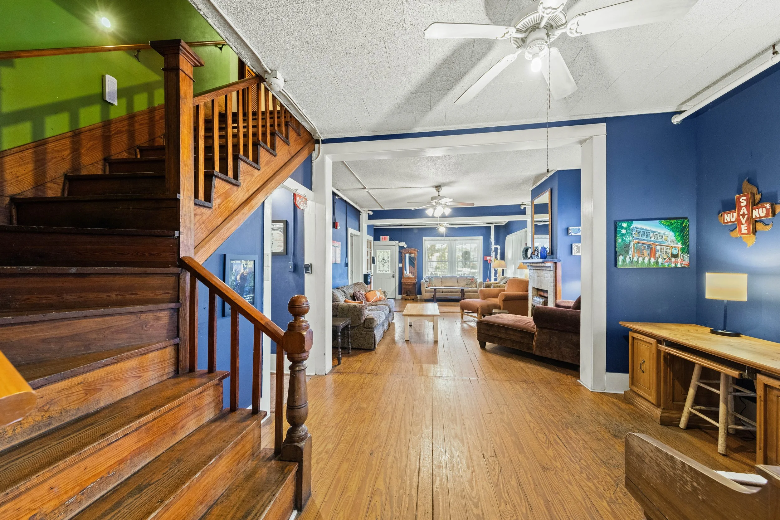 Interior of a cozy living room with blue and green walls, wooden stairs, hardwood floors, multiple sofas, a fireplace, ceiling fans, and decorations including paintings and wall hangings.