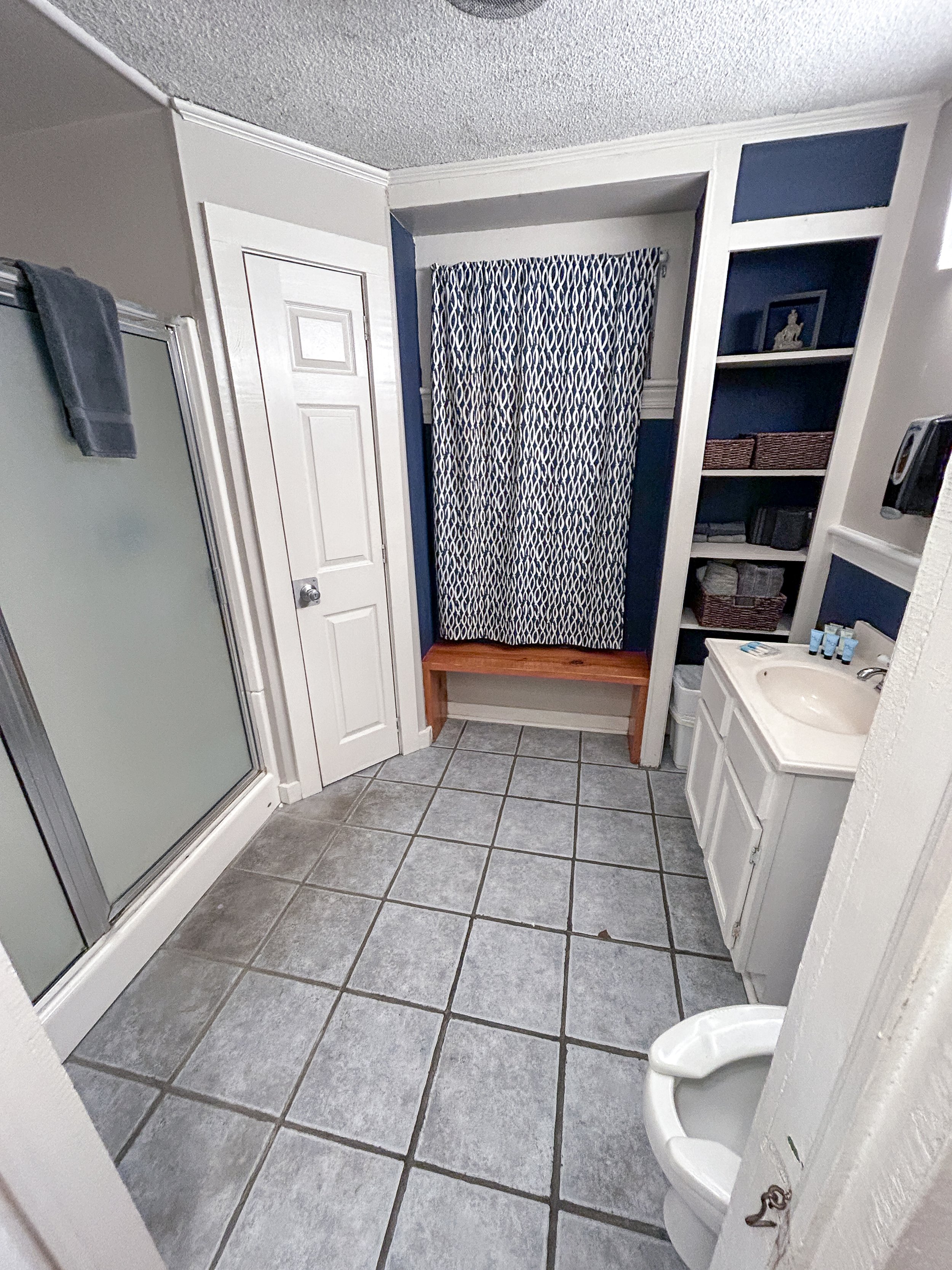 A bathroom with a shower on the left, a closed white closet door, a navy and white patterned curtain, with built-in shelves on the right, a small white sink, and a water closet (toilet) without a seat, all on gray tiled flooring.