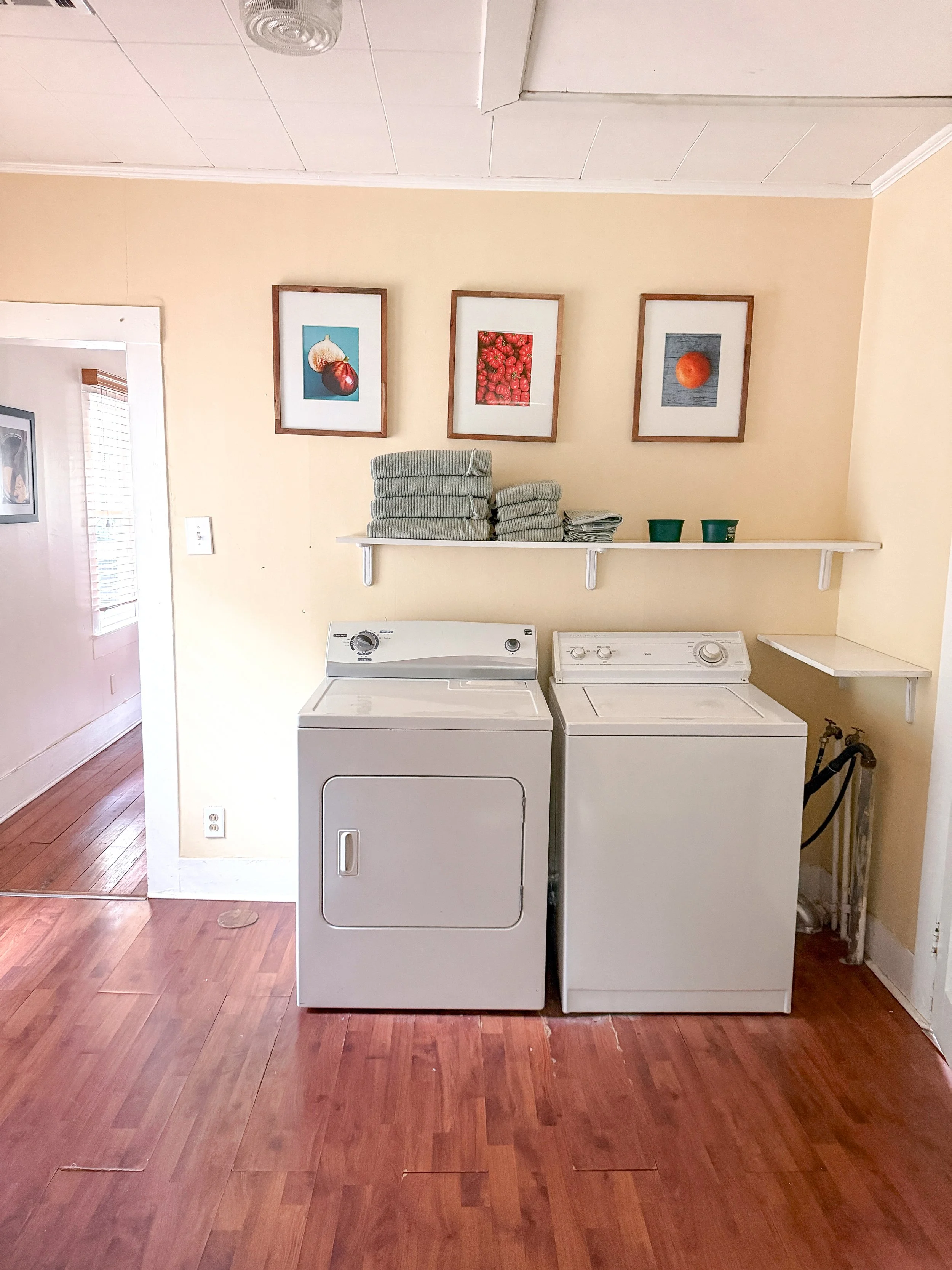 A laundry room with a stacked washer and dryer, a white shelf holding folded towels and two green cups, three framed pictures of fruits on the wall, and wooden flooring.