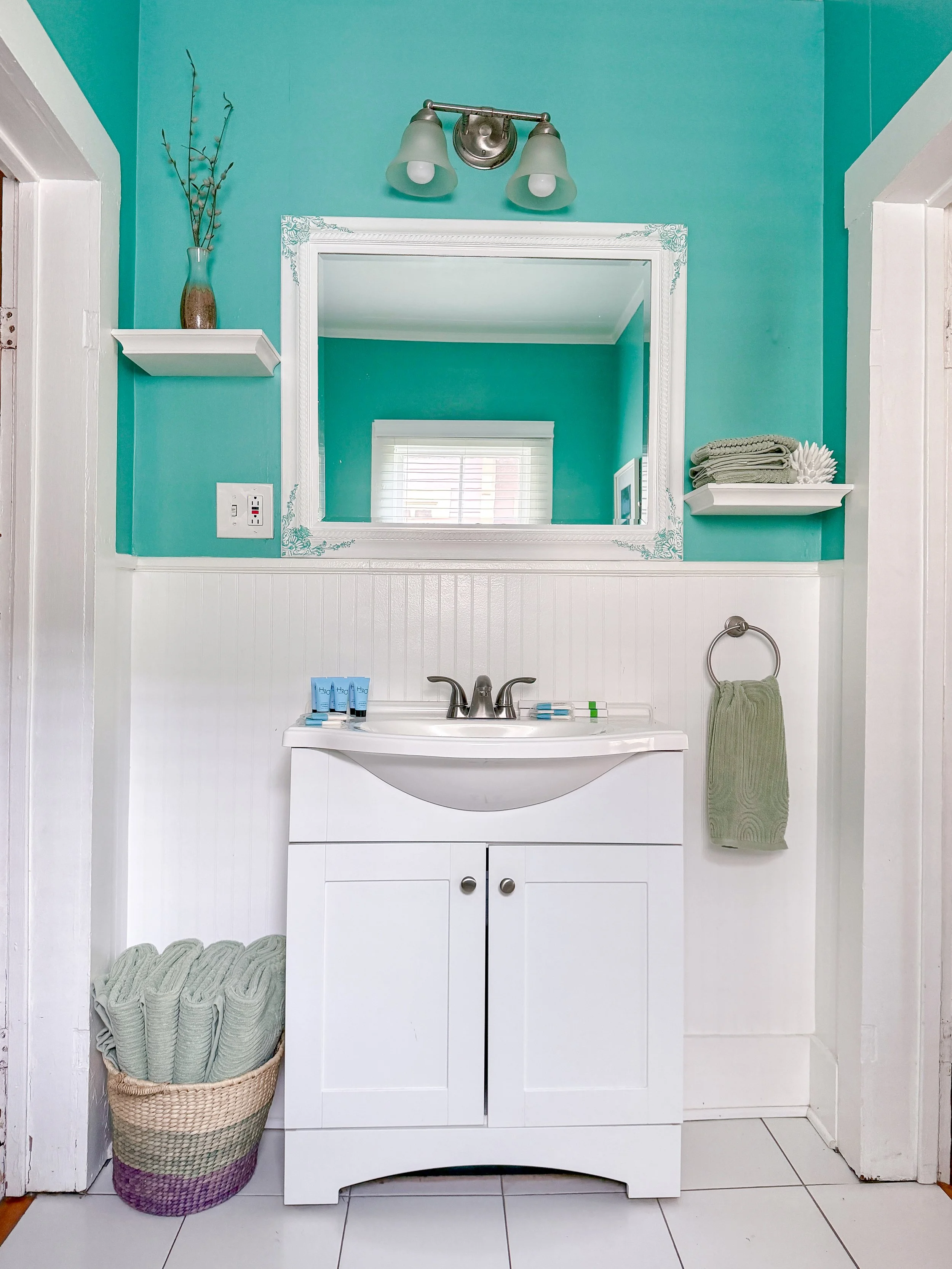 Bathroom with teal upper wall and white lower beadboard paneling, white sink vanity with a mirror reflecting a window, wall-mounted light fixture, folded towels, and decorative touches, including a basket of rolled towels on the floor.