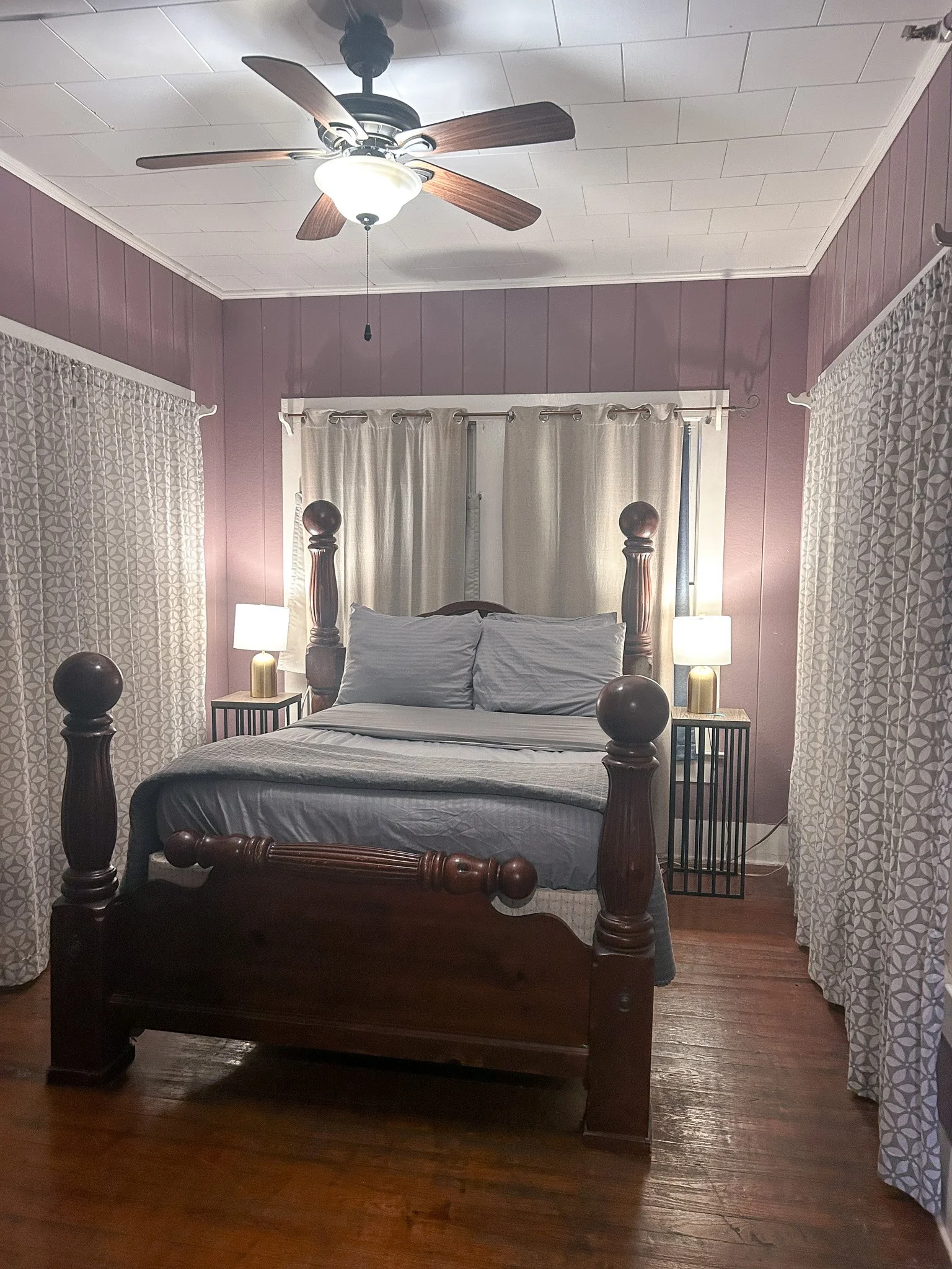 A bedroom with a wooden four-poster bed, white bedding, and two nightstands with lamps. Purple walls, patterned curtains, wood flooring, and a ceiling fan.