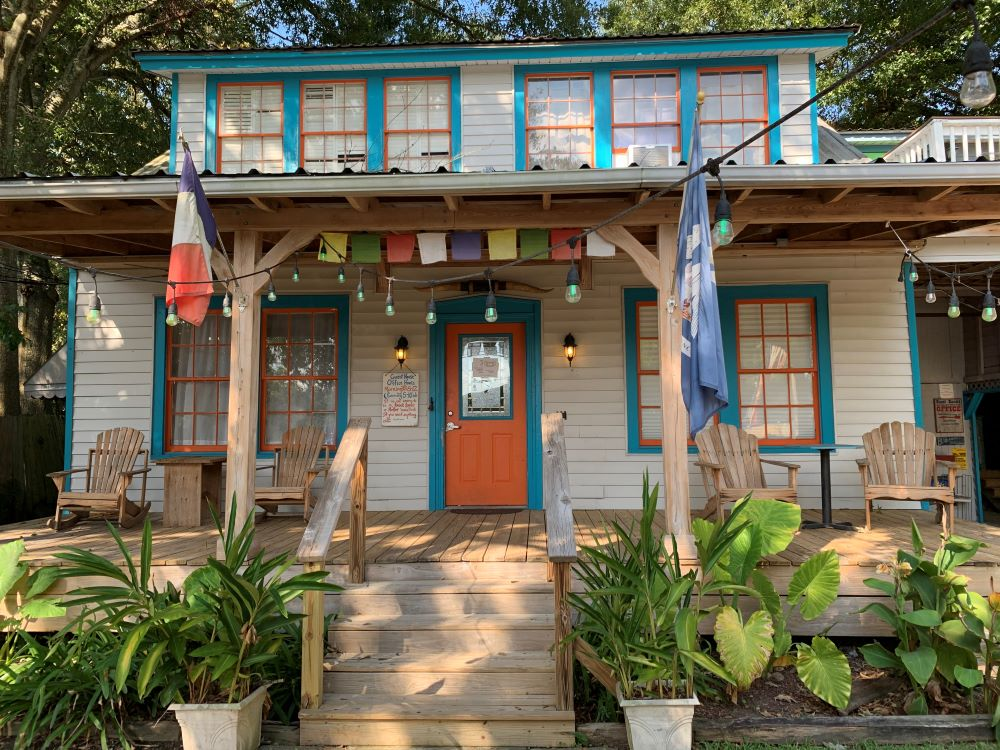 Front view of a two-story wooden building with outdoor seating, string lights, potted plants, and flags, with a colorful paint scheme and large windows.