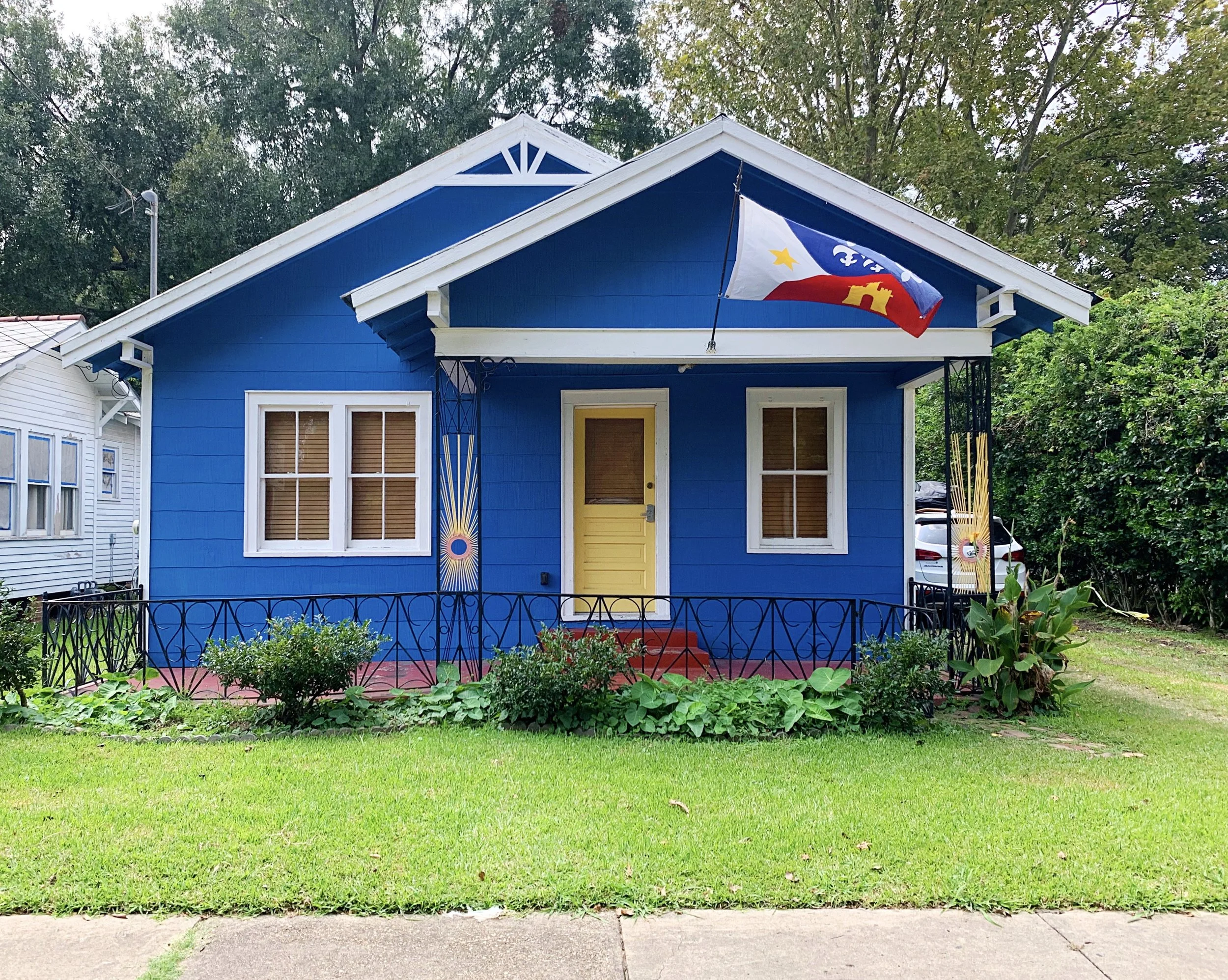 Blue house with yellow door, white trim, a porch with a black railing, and a flag hanging from the roof. Green lawn and bushes in front, with trees in the background.