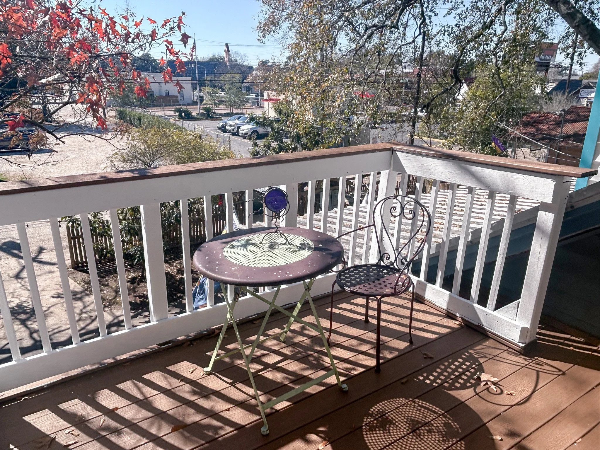 A small balcony with a round metal table and a metal chair overlooking a parking lot and trees with autumn leaves.