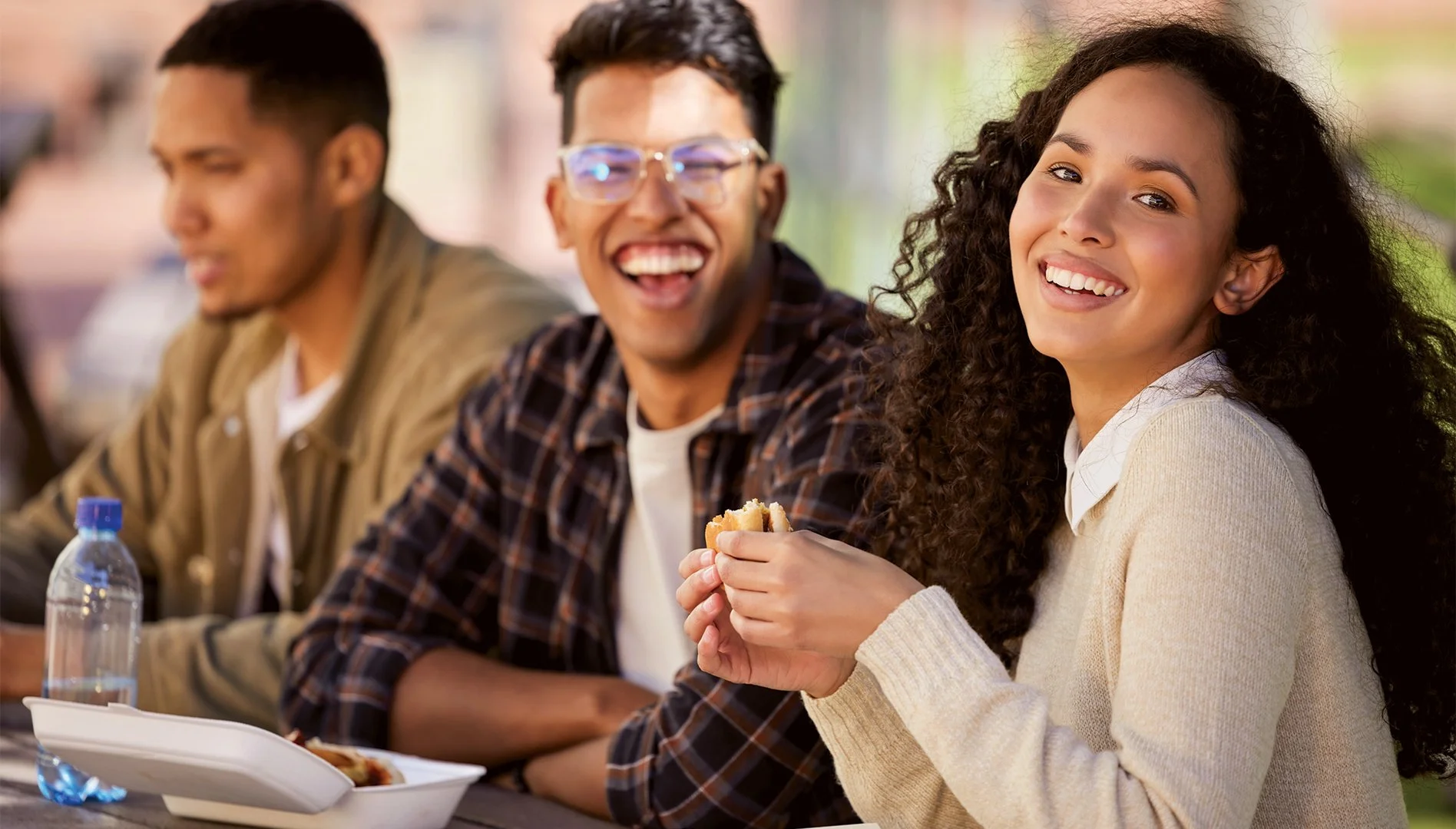 Grupo de jovens adultos sorrindo, sentado na mesa ao ar livre, com comida e bebida, em ambiente ao ar livre, durante o dia.