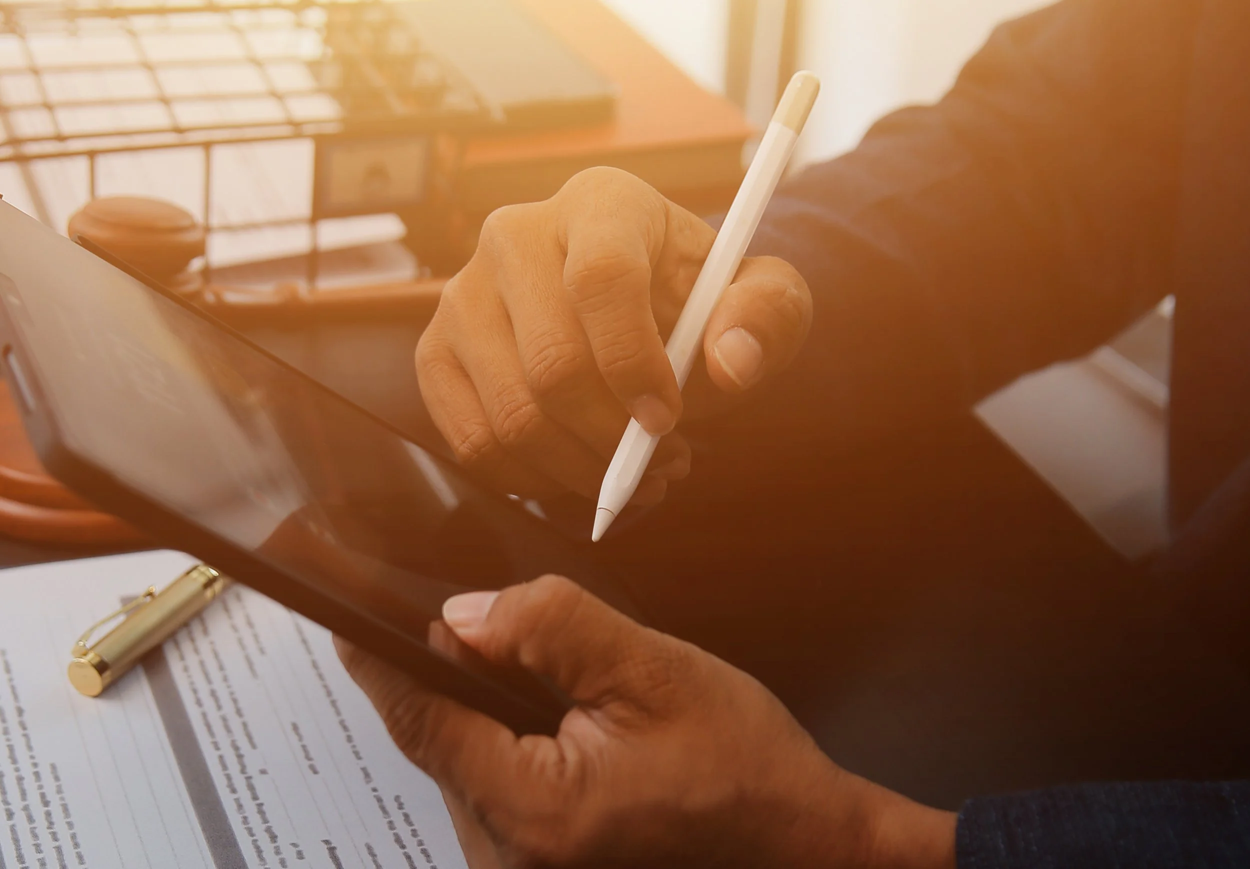 Pessoa usando um iPad e uma caneta digital para fazer anotações ou desenhar, com documentos e uma caneta de alta qualidade na mesa.