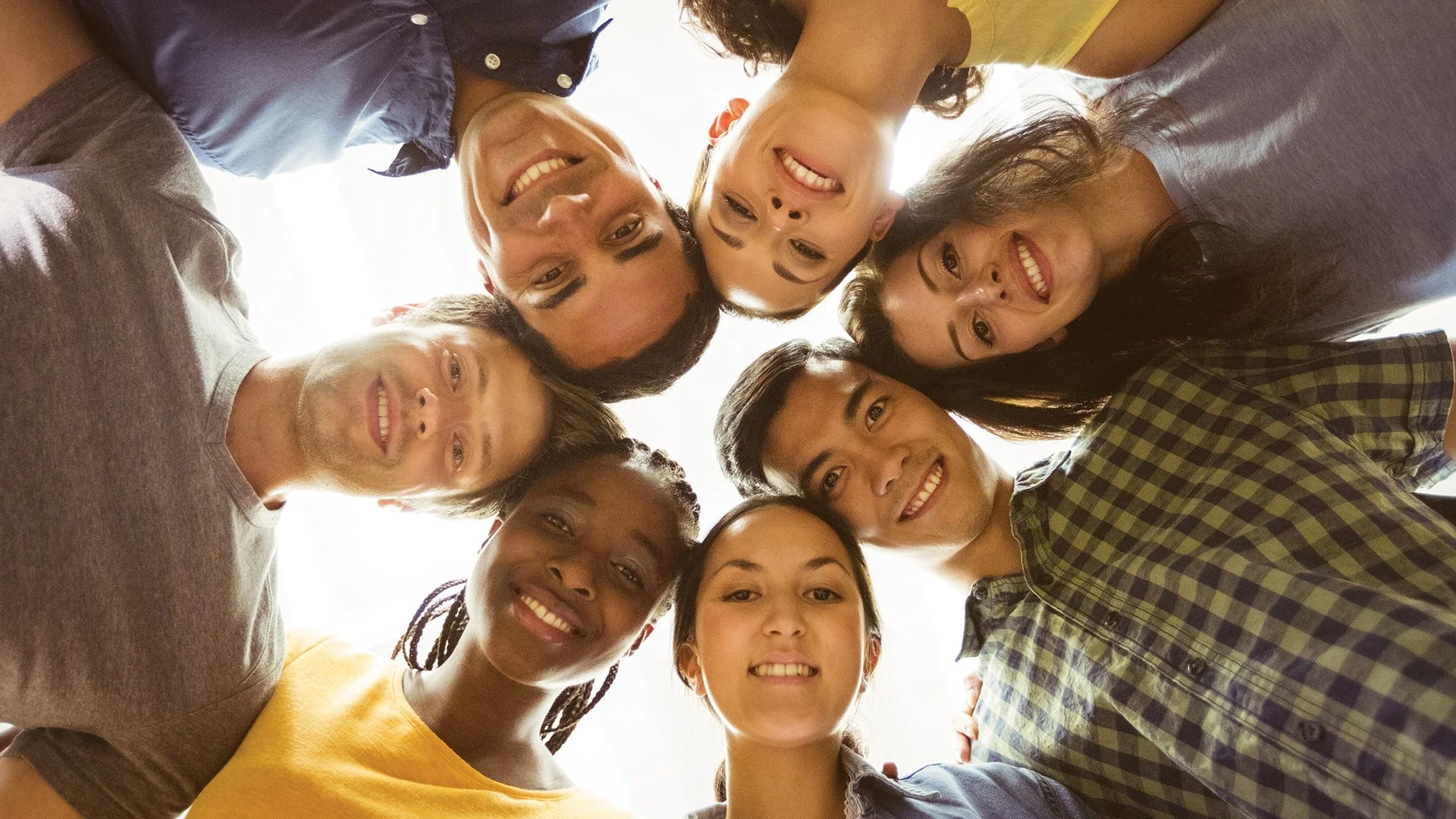 Grupo de pessoas diversas olhando para a câmera, sorrindo, formando um círculo visto de baixo, laje ensolarada ao fundo.
