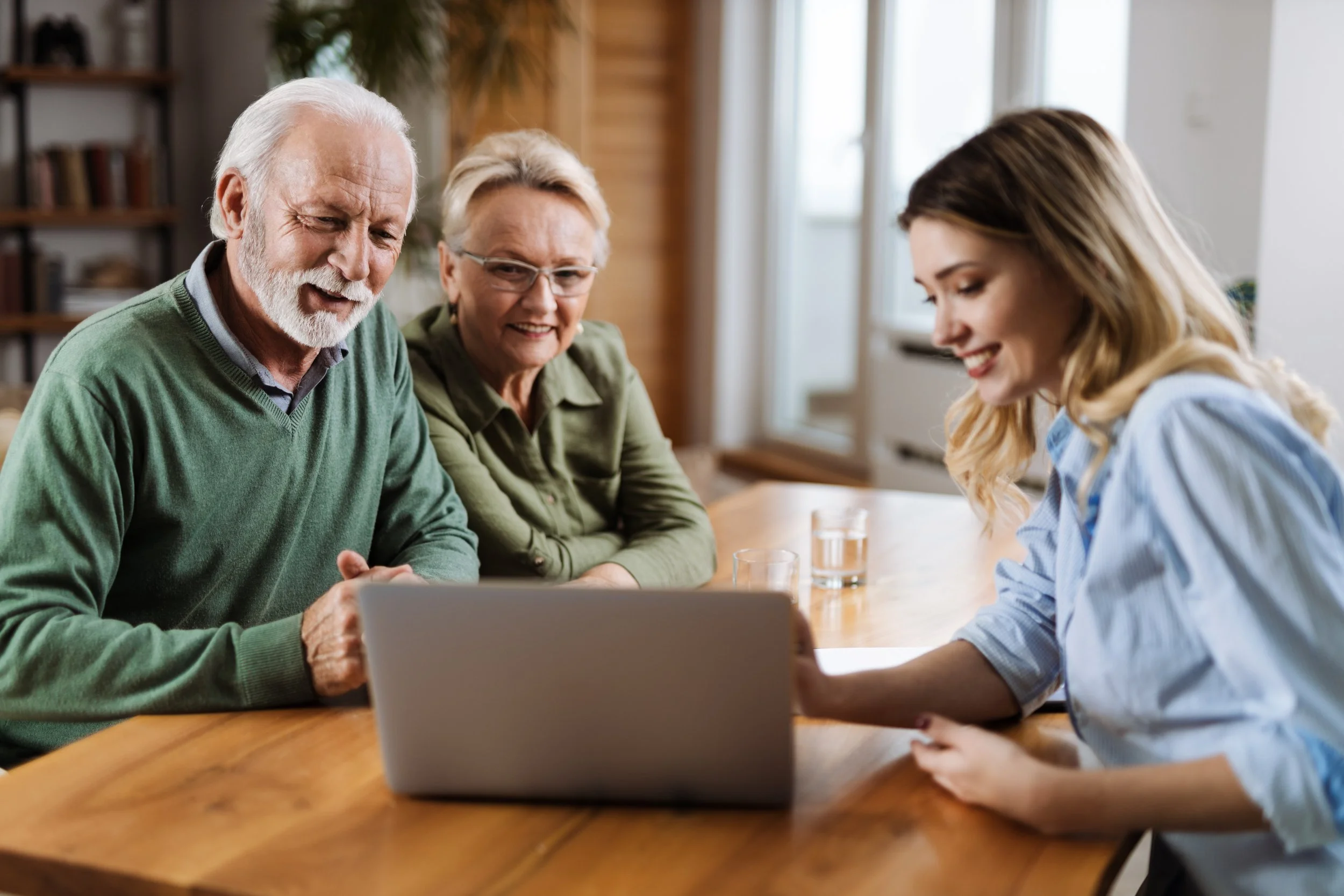Three elderly people and a young woman sit at a wooden table looking at a laptop, smiling and engaged in a video call in a cozy, well-lit room.