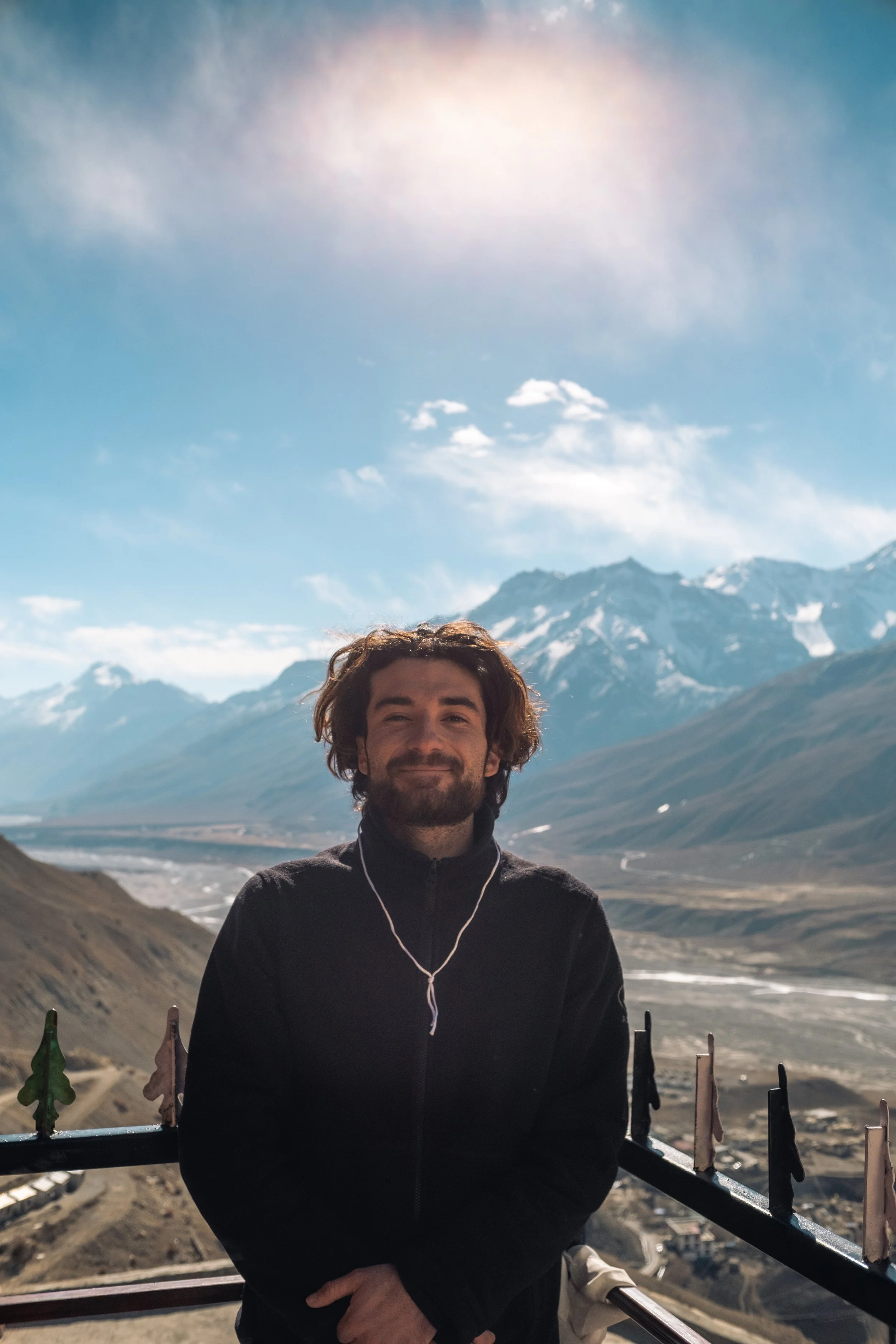 Un homme avec des cheveux bouclés et une barbe se tient devant un paysage montagneux, portant un parka noir et des écouteurs.