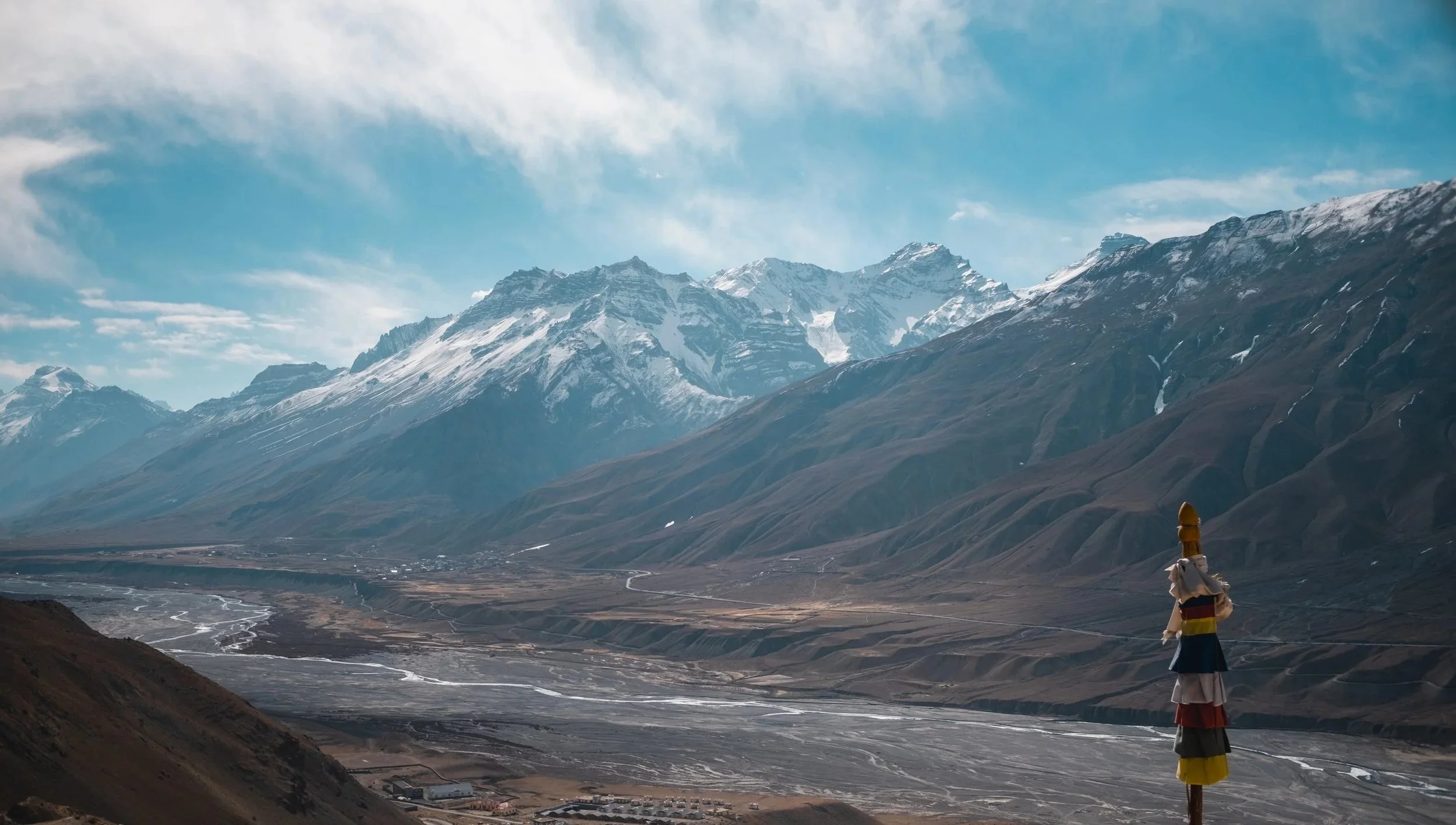 Mountainous landscape with snow-capped peaks, valleys, and a decorative fabric pole in the foreground.