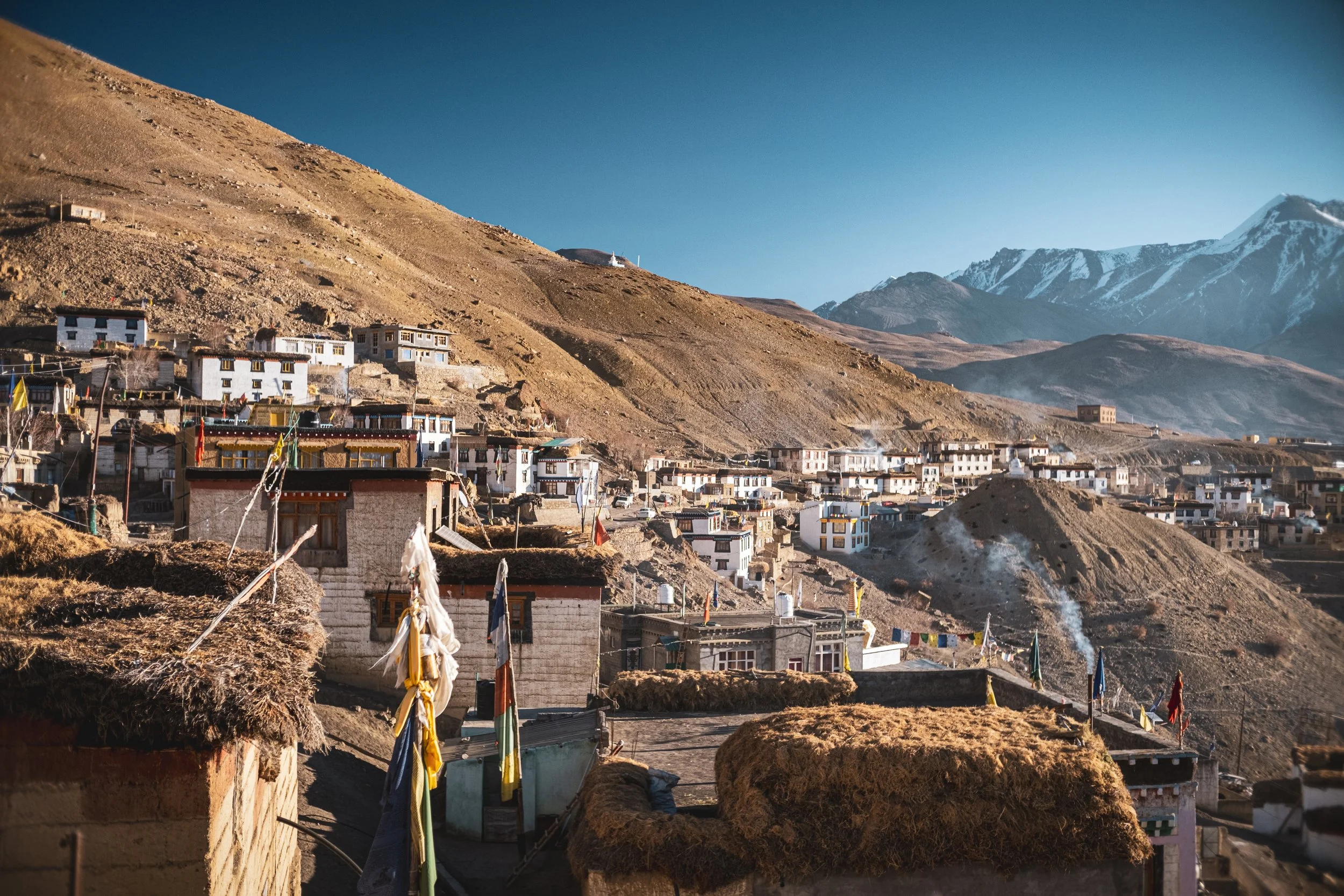 Vue panoramique d'un village himalayen avec des maisons en pierre et en bois, entouré de montagnes enneigées au loin, sous un ciel bleu clair.