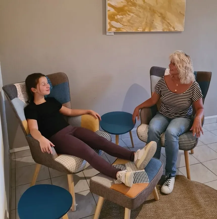 A young girl and an older woman are sitting in armchairs, engaging in a conversation. The girl has dark hair and is wearing a black shirt and maroon pants, with her legs stretched out and resting on a footstool. The woman has curly blonde hair and is wearing a striped shirt and jeans. They are sitting in a cozy, modern room with light-colored walls, abstract artwork, small blue side tables, and a rug on the tiled floor.