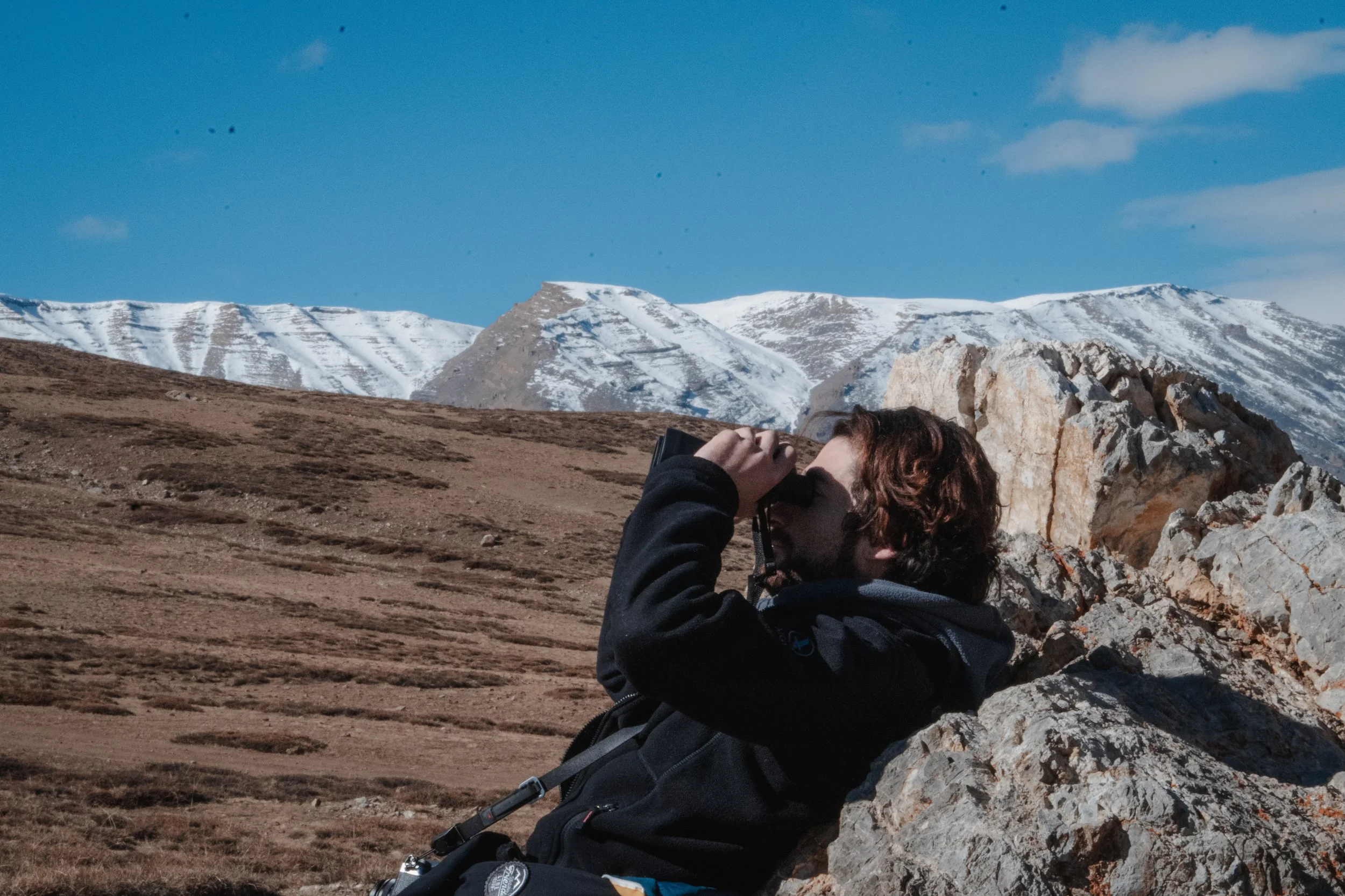 Un homme avec des cheveux marrons foncés, portant un sweat noir, regarde à travers des jumelles dans un paysage de montagne avec des sommets enneigés sous un ciel bleu.