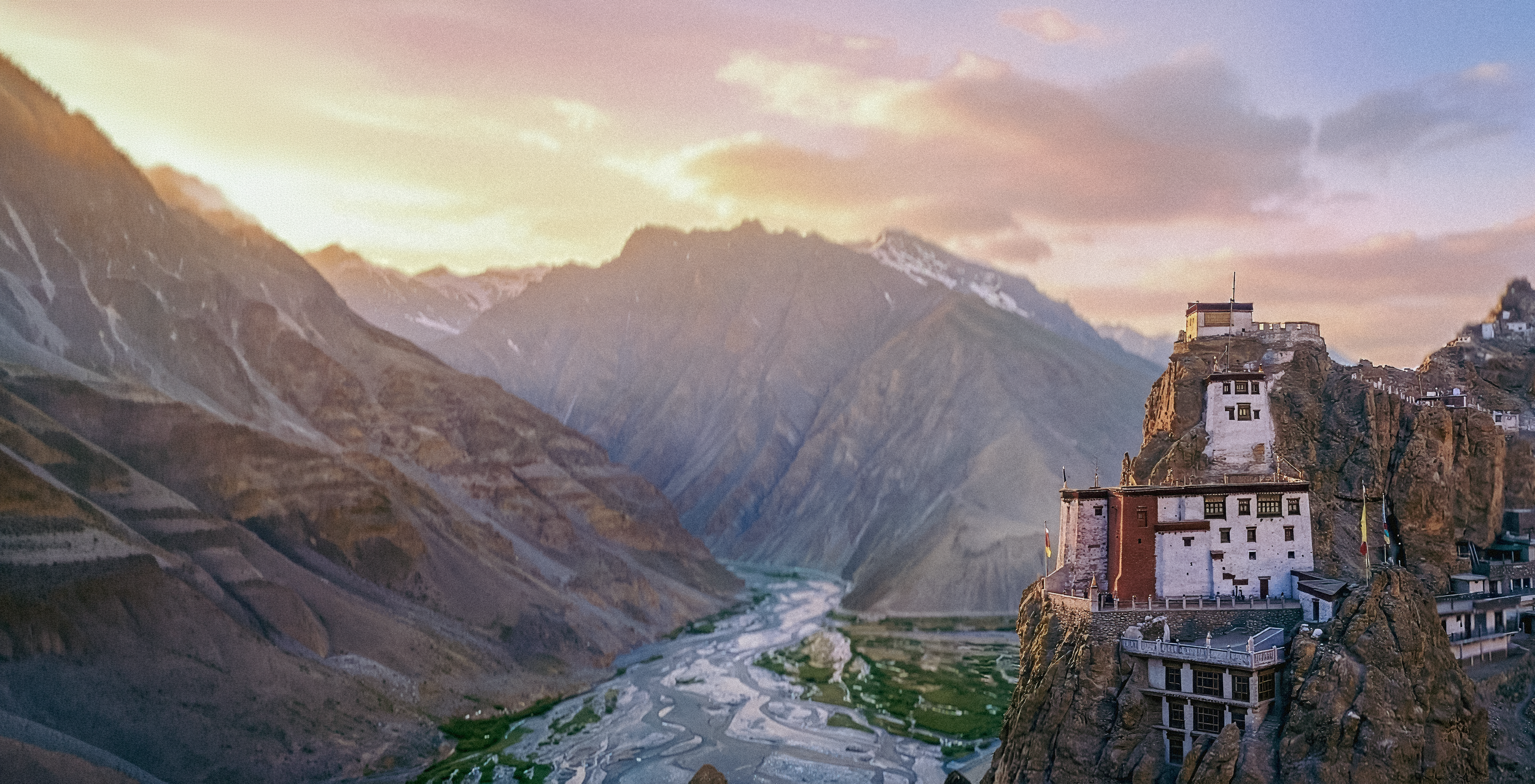 Vue panoramique de montagnes avec un château construit sur une falaise sur la droite, et une vallée avec une rivière au fond.