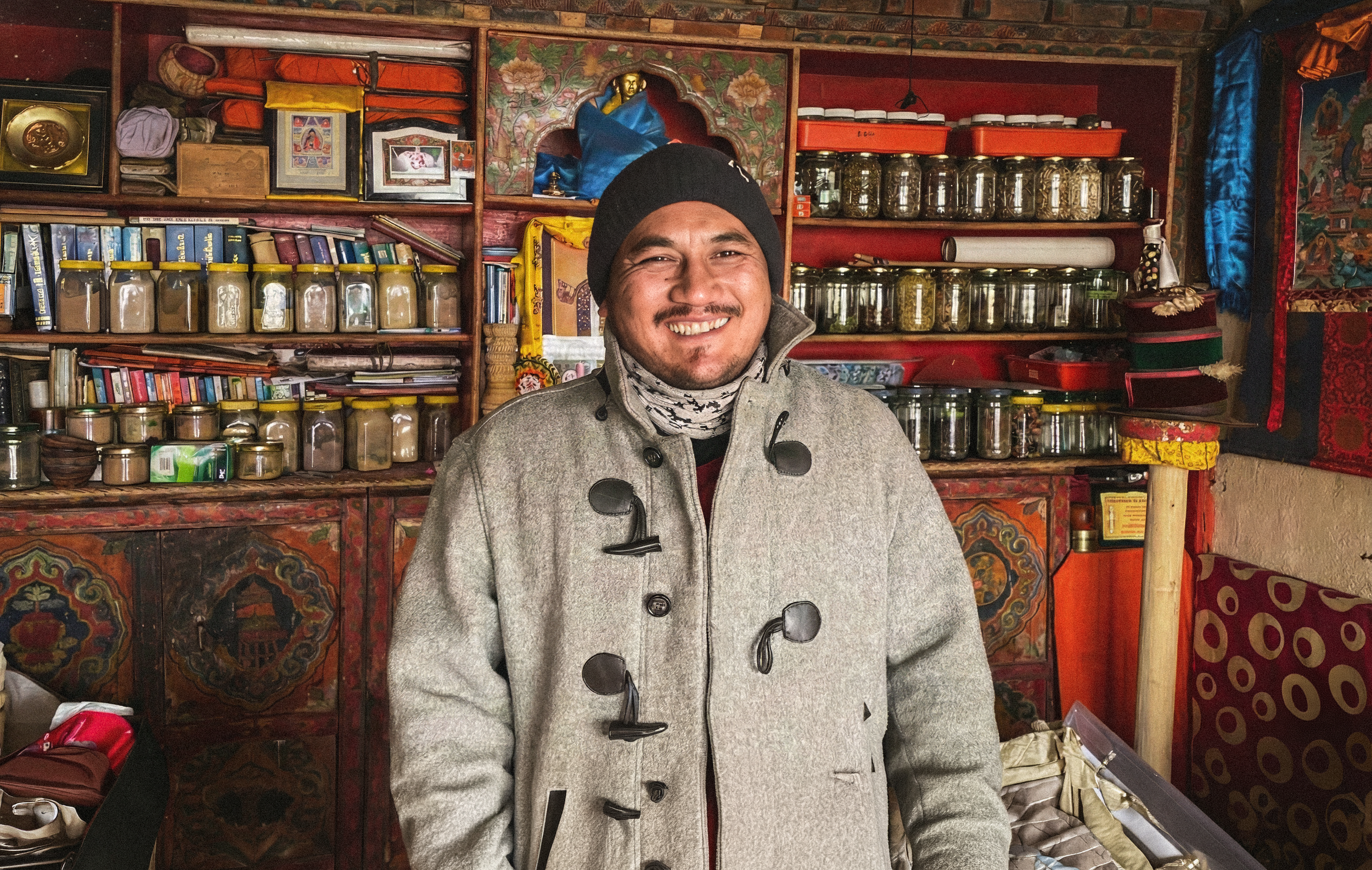 Norbu, A smiling man wearing a gray coat, black beanie, and a patterned white scarf, standing in a shop or market filled with colorful jars and handmade items.