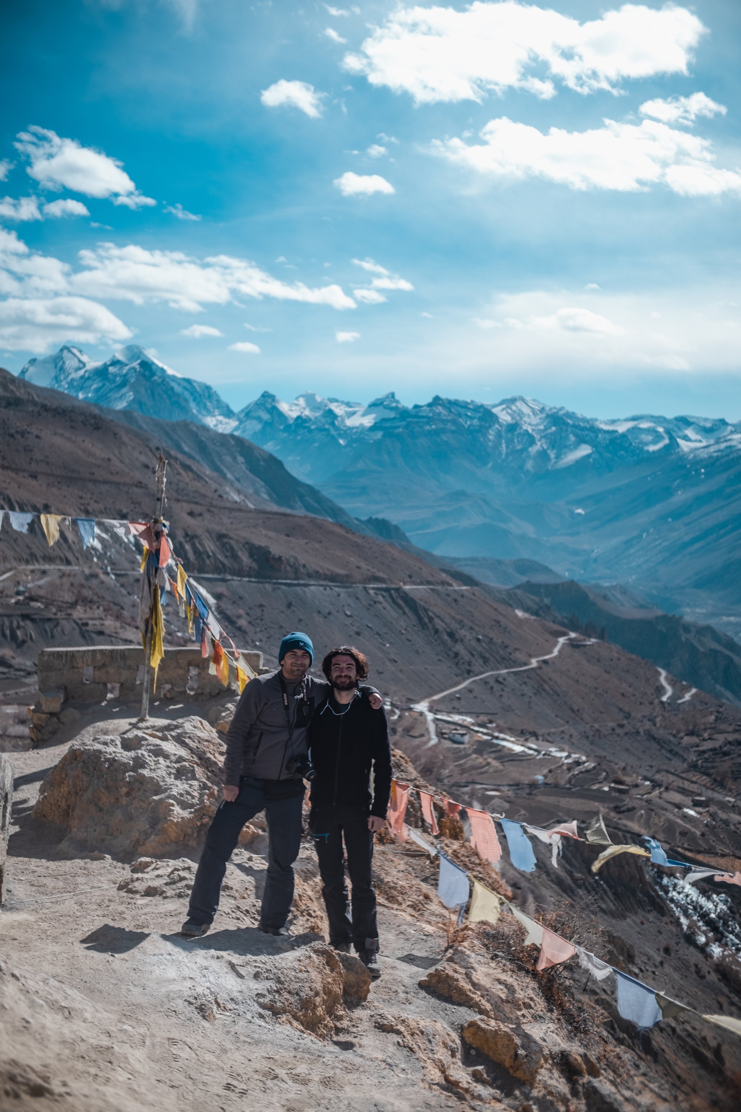 Deux hommes posent ensemble sur un sentier de montagne avec des montagnes enneigées en arrière-plan et des drapeaux de prière tibétains suspendus