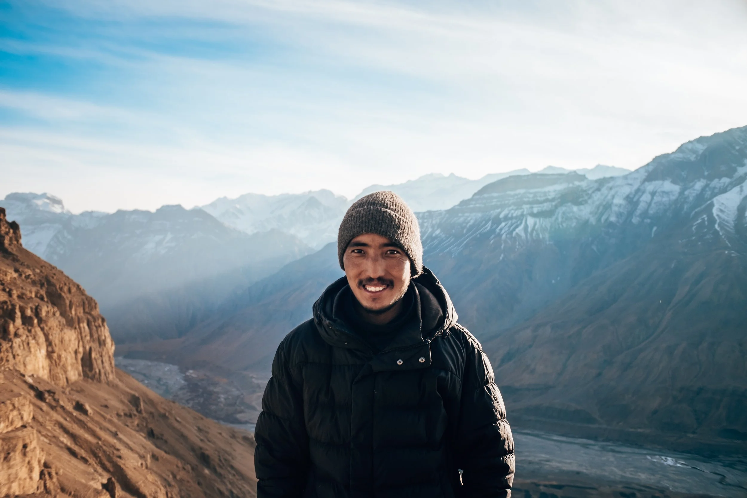 Un homme souriant en vêtements d'extérieur dans un paysage de montagnes enneigées et de vallées.
