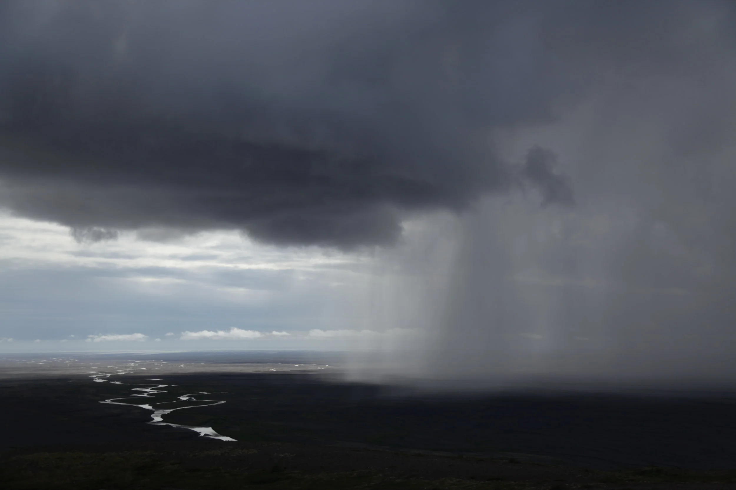 Dark storm clouds over a flat landscape with a heavy rain shower visible from the clouds to the ground, and a winding river below.