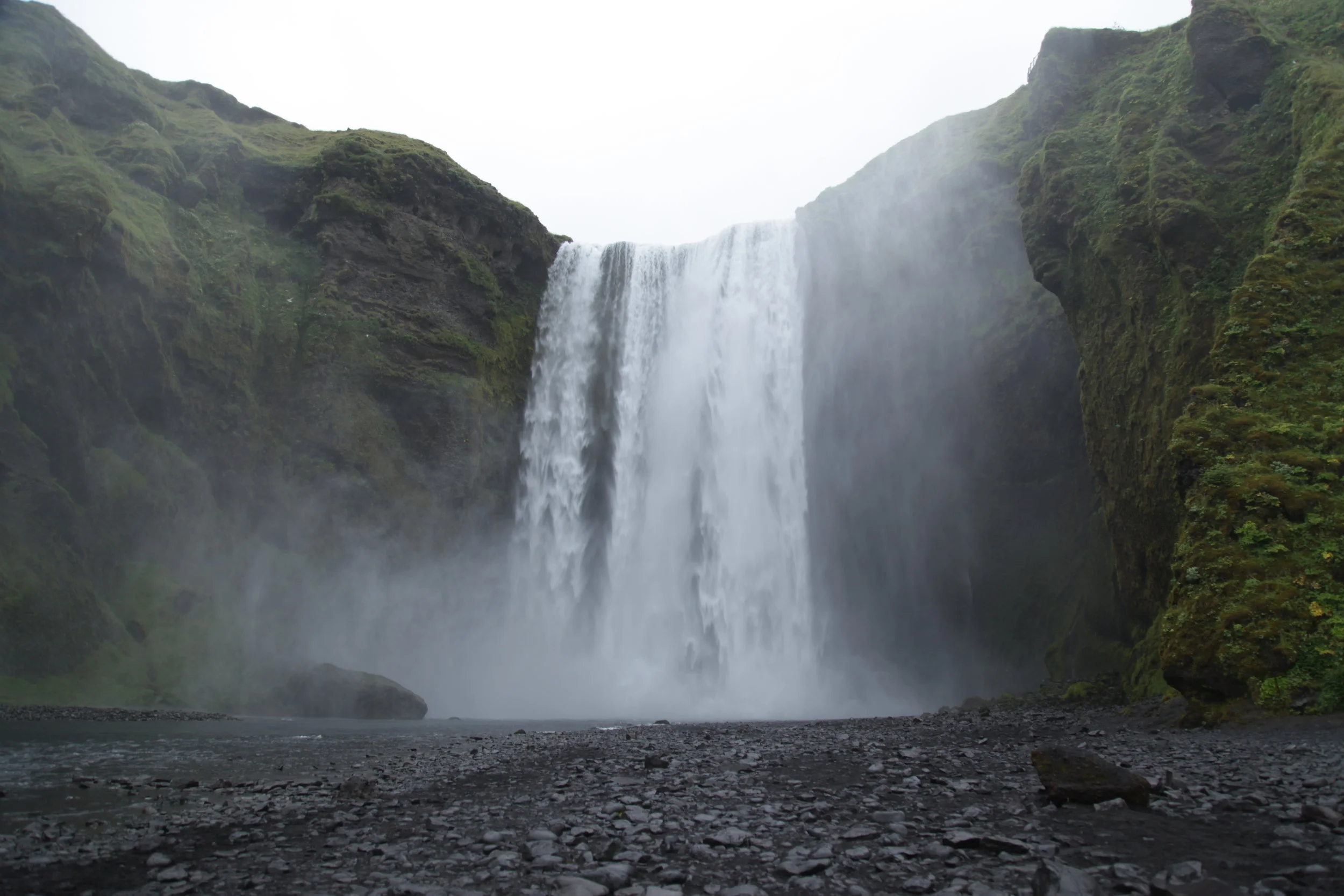 A large waterfall cascading down mossy green cliffs into a rocky river below with mist rising around the base.