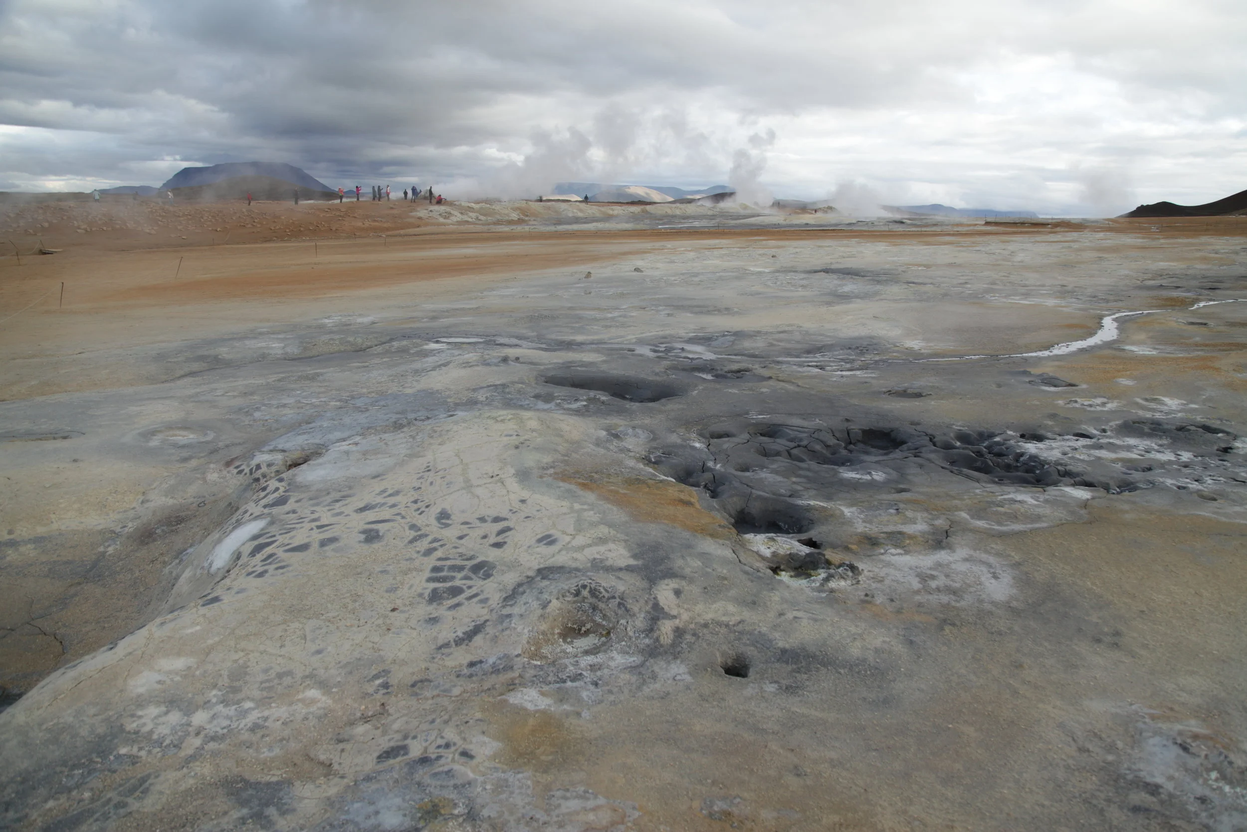 Geothermal area with steaming vents, cracked earth, and sulfur deposits under cloudy sky, with people in the background on a boardwalk.