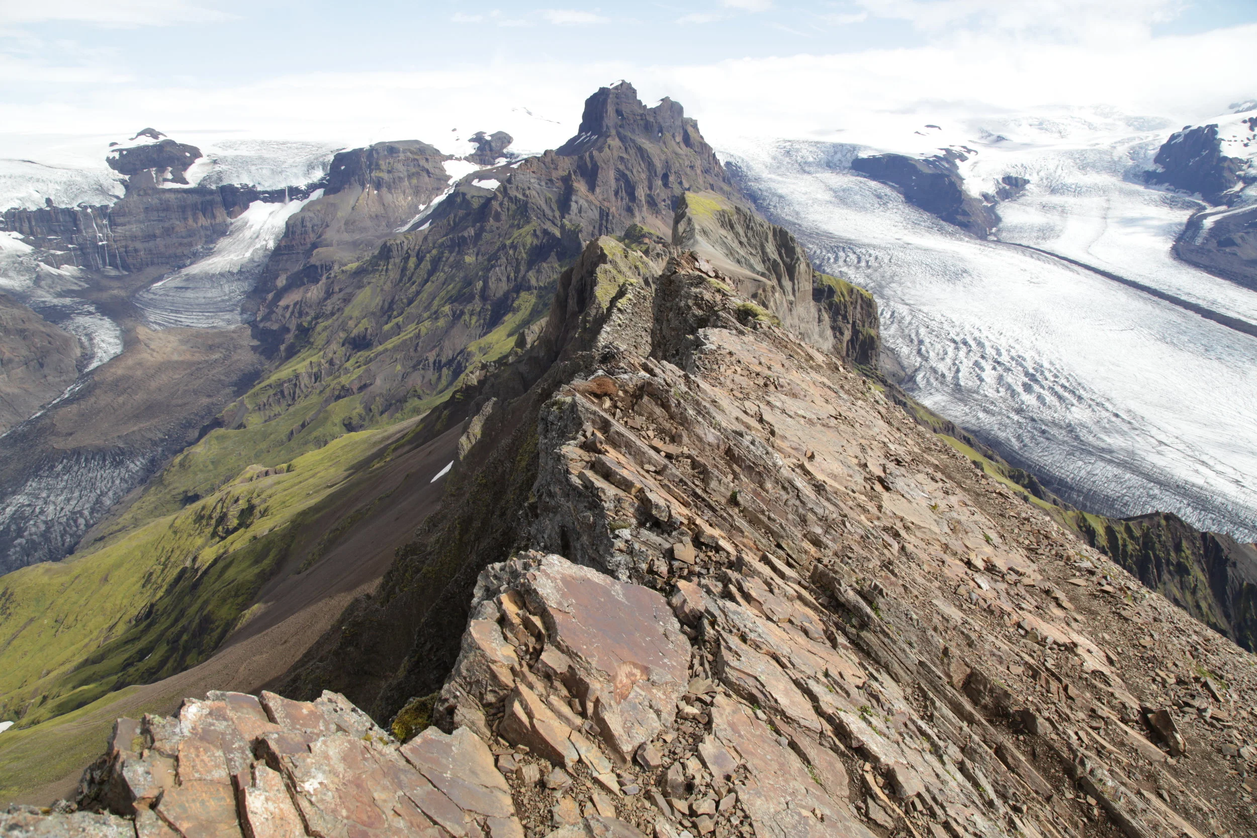 Mountain ridge with rocky terrain leading to snow-capped peaks and glaciers in background.