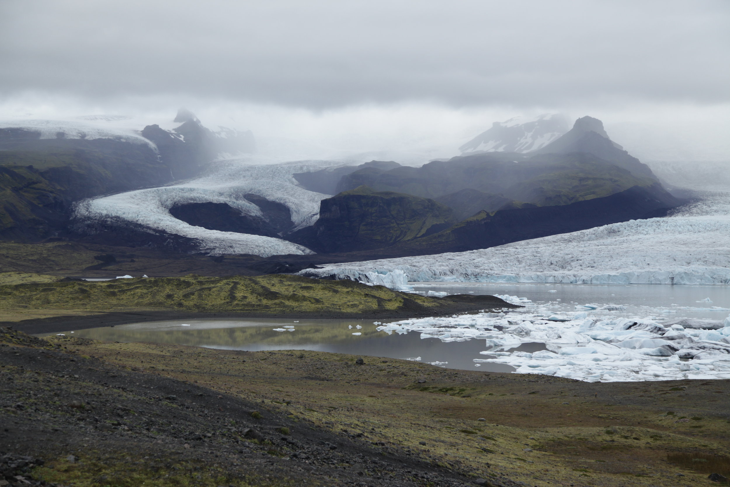 Icelandic landscape with glaciers, mountains, and a partially frozen lake under cloudy sky.