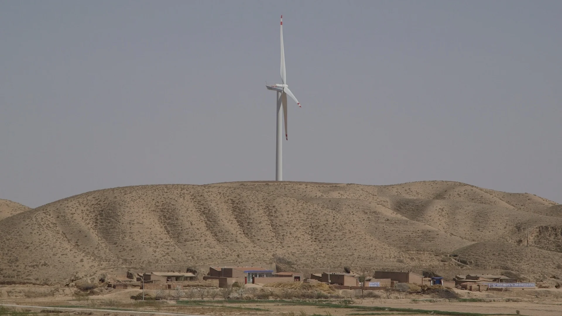 A wind turbine on a hillside with a small cluster of buildings below, set in a desert landscape with sandy, barren hills and clear sky.